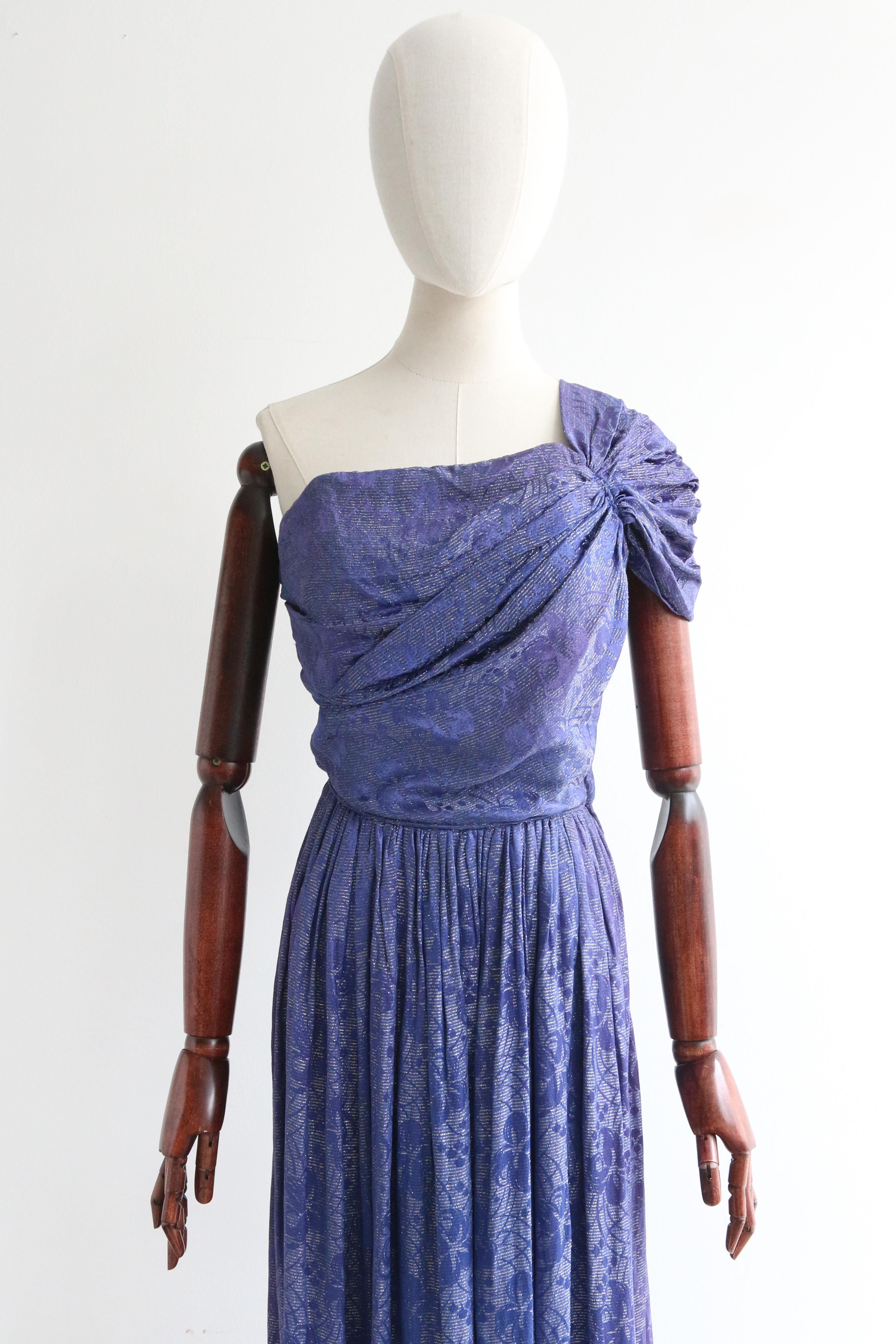 Este fascinante vestido de brocado de seda azul noche de los años 30, con un acento de hilo lamé plateado, es una pieza rara de contemplar

El escote recto del vestido se realza con un maravilloso tirante único, elegantemente plisado y fruncido, que