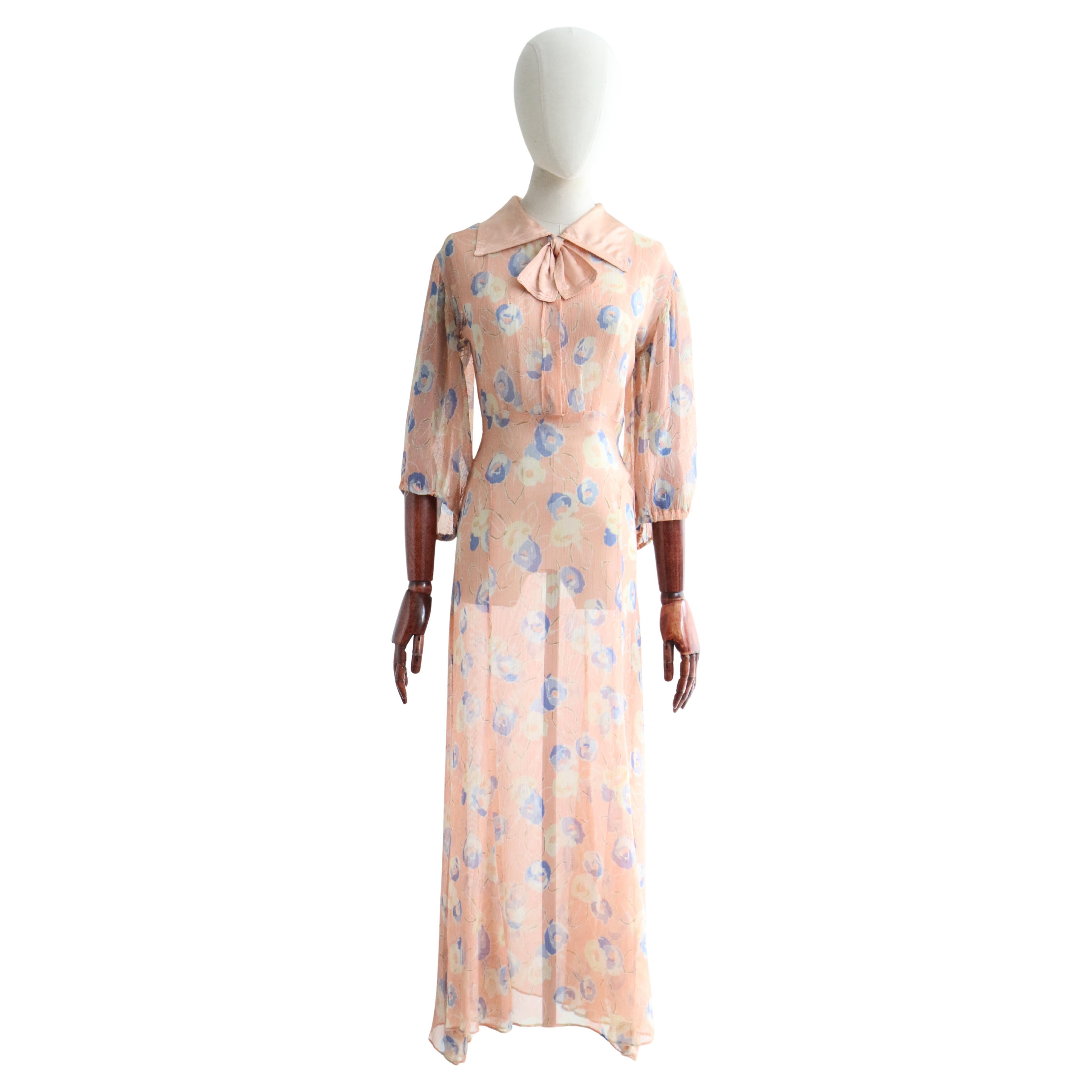 This romantic 1930's dress, in a soft floral pattern in shades of blue and cream, with muted olive green accents, set against a blush pink background, is just the piece to add to your seasonal wardrobe.  
  
The V shaped neckline of the dress is