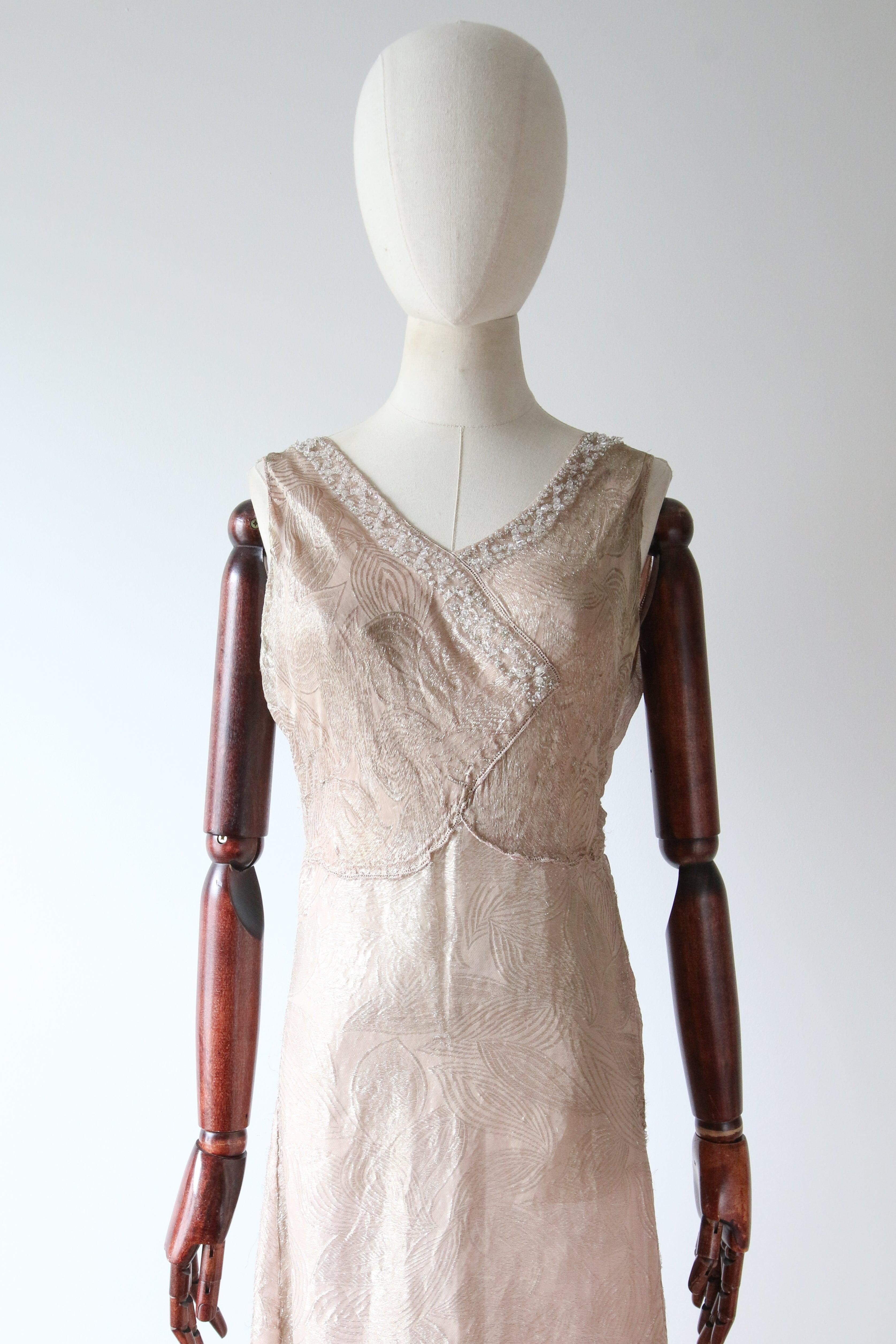 This truly enchanting 1930's blush silk and gold lamé dress, with a glistening pattern of leaves, finished with glass beaded embellishments along the neckline, is a rare piece to behold.  
The V shaped neckline is framed by wide shoulder straps and