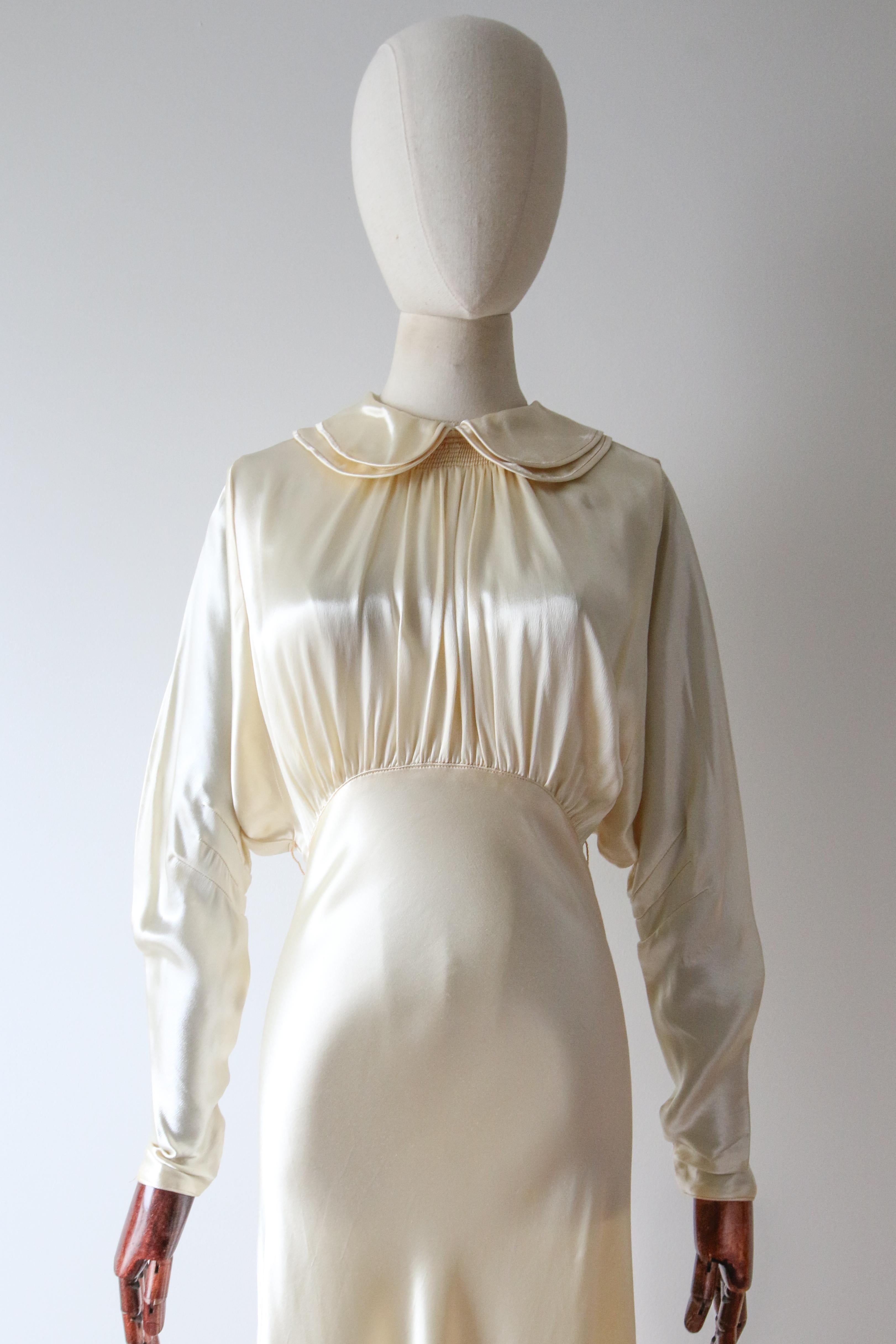 Réalisée dans un incroyable satin crème et dotée de délicats boutons en strass argenté, cette magnifique robe à coupe en biais des années 1930 est un véritable morceau d'histoire de la mode. 

L'encolure arrondie est encadrée par un doux col Peters