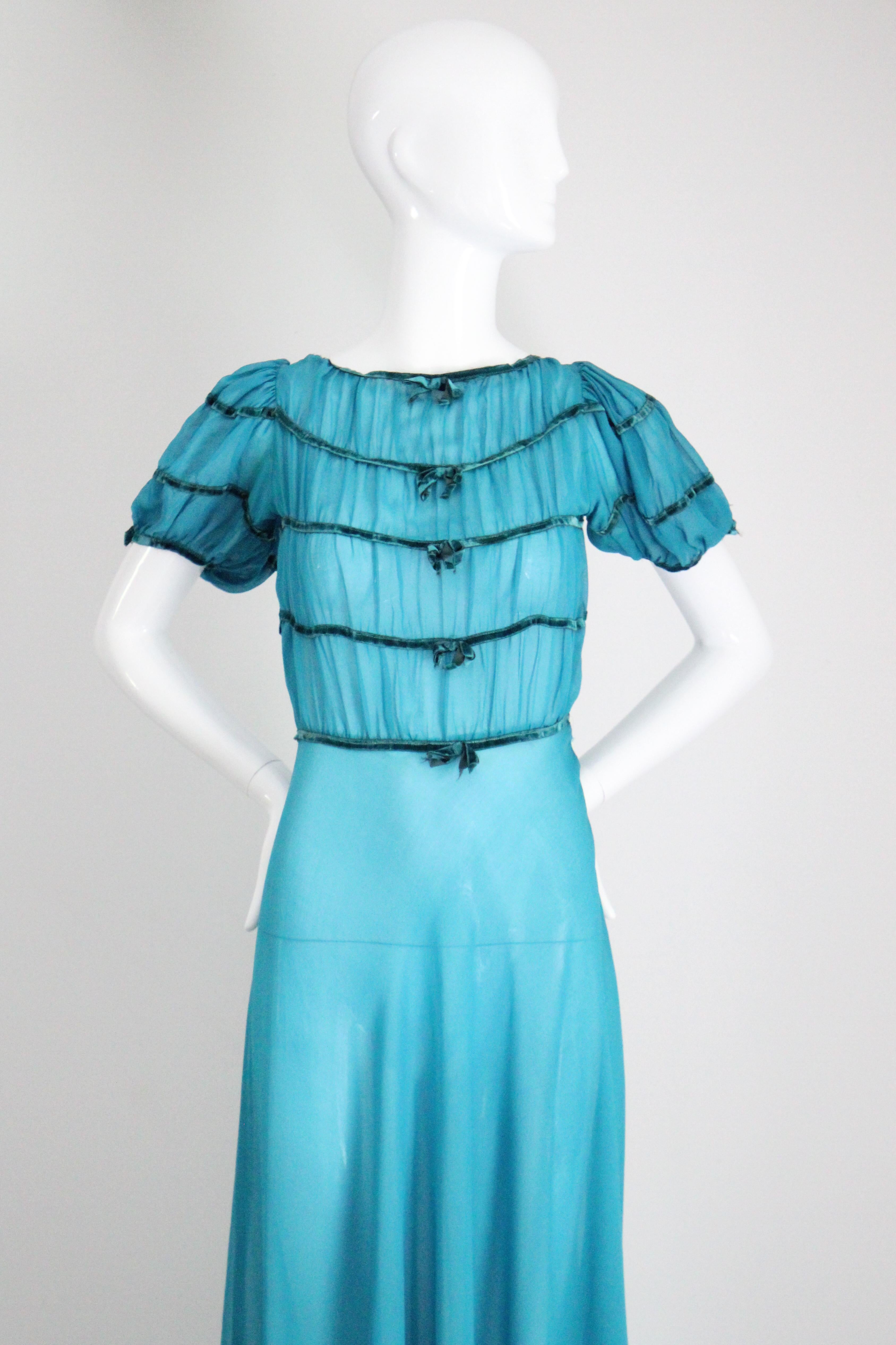 Réalisée dans la plus incroyable nuance de mousseline de soie bleu turquoise, rehaussée de rubans de velours sarcelle, cette robe vintage originale des années 1930 est une pièce rare à contempler. 

L'encolure arrondie est encadrée par une bordure