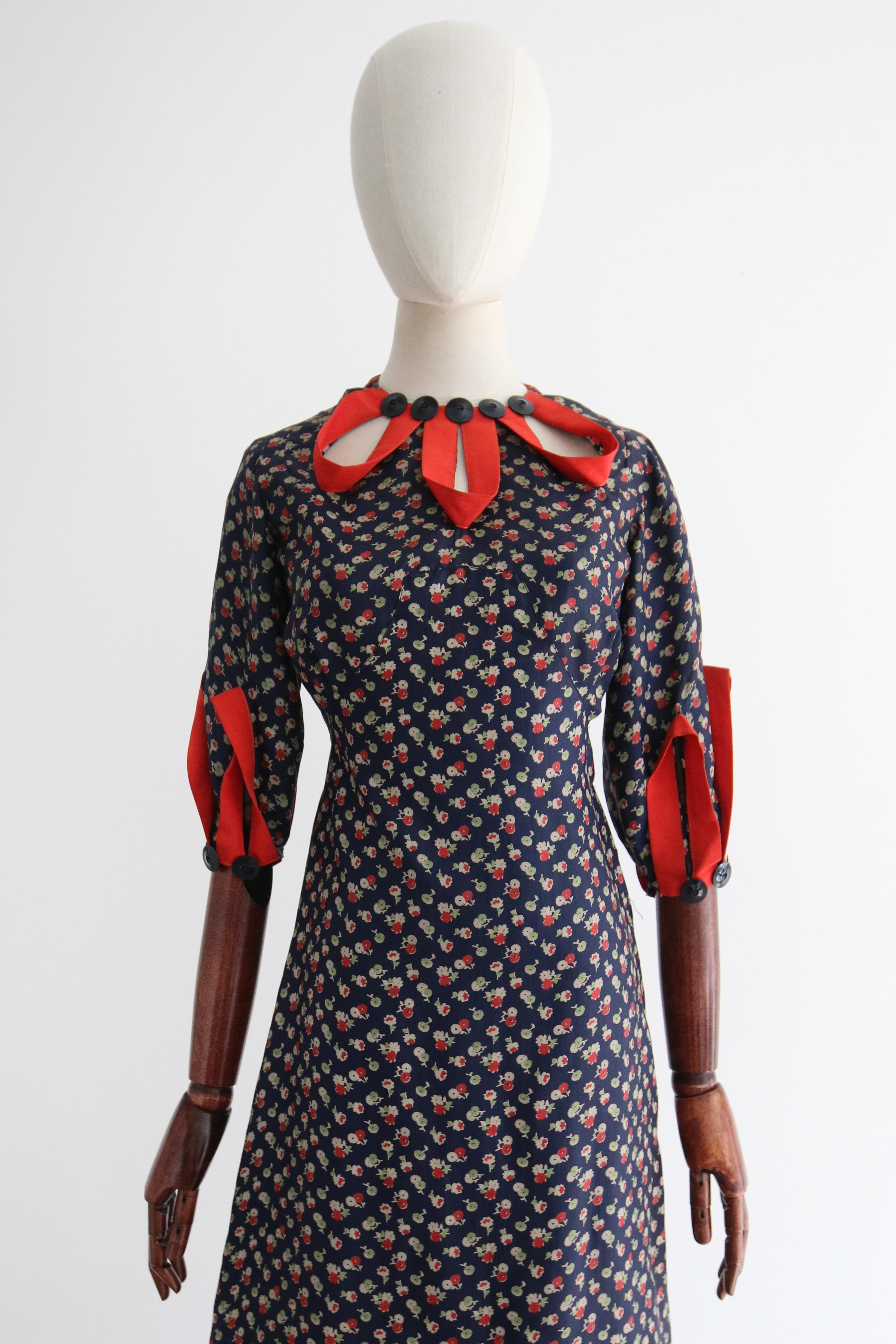 A marvel of its time, this original 1930's blue twill silk dress with a floral pattern in shades of grey, green and red, finished with red faille ribbon details and navy blue buttons, is a rare piece to behold. 

The rounded neckline of the dress is