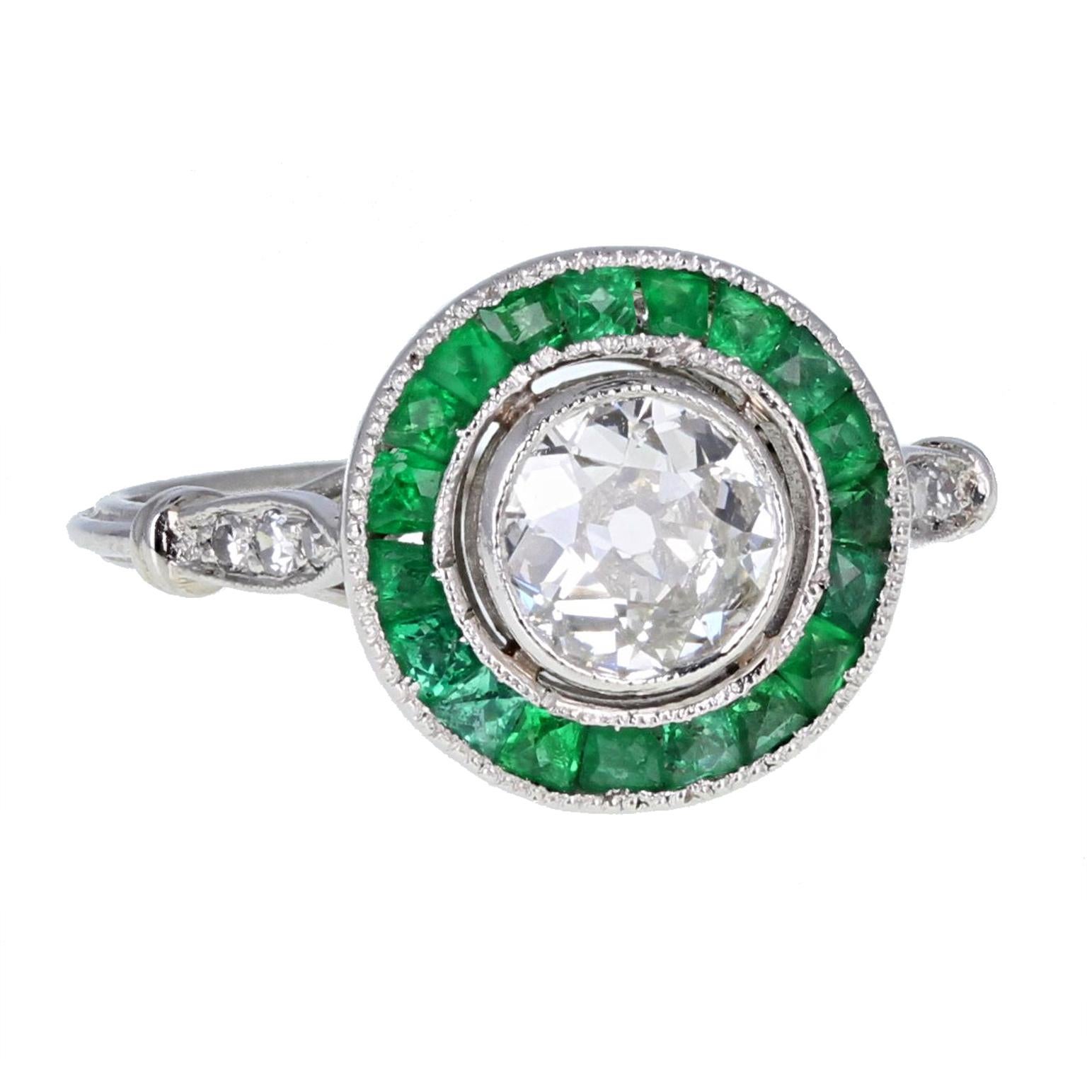 An exquisitely crafted 'target' ring. A central old cushion-cut diamond of approximately 1 carat set in a bezel setting with millegrain detailing. Surrounded by a halo of beautiful green calibré-cut emeralds to form a target style cluster.