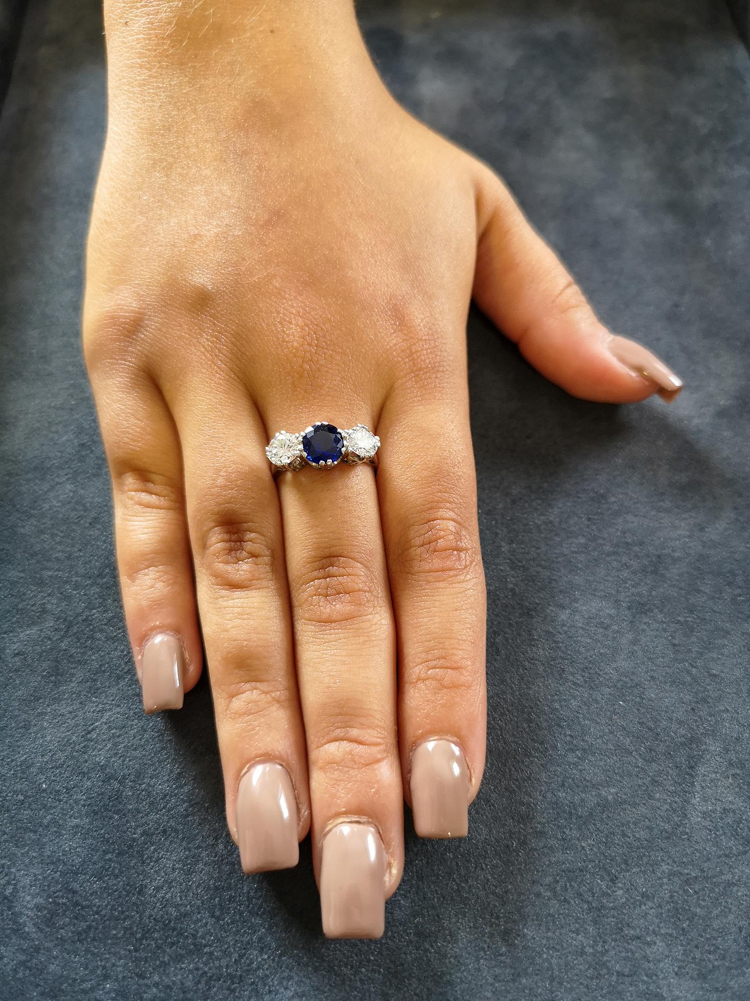 A traditional platinum basket-style setting with double-claws adorned with a central cushion-shaped inky-blue sapphire of approximately 1.00 carat, flanked on either side by a single brilliant-cut diamond, each approximately 0.50 of a carat, H