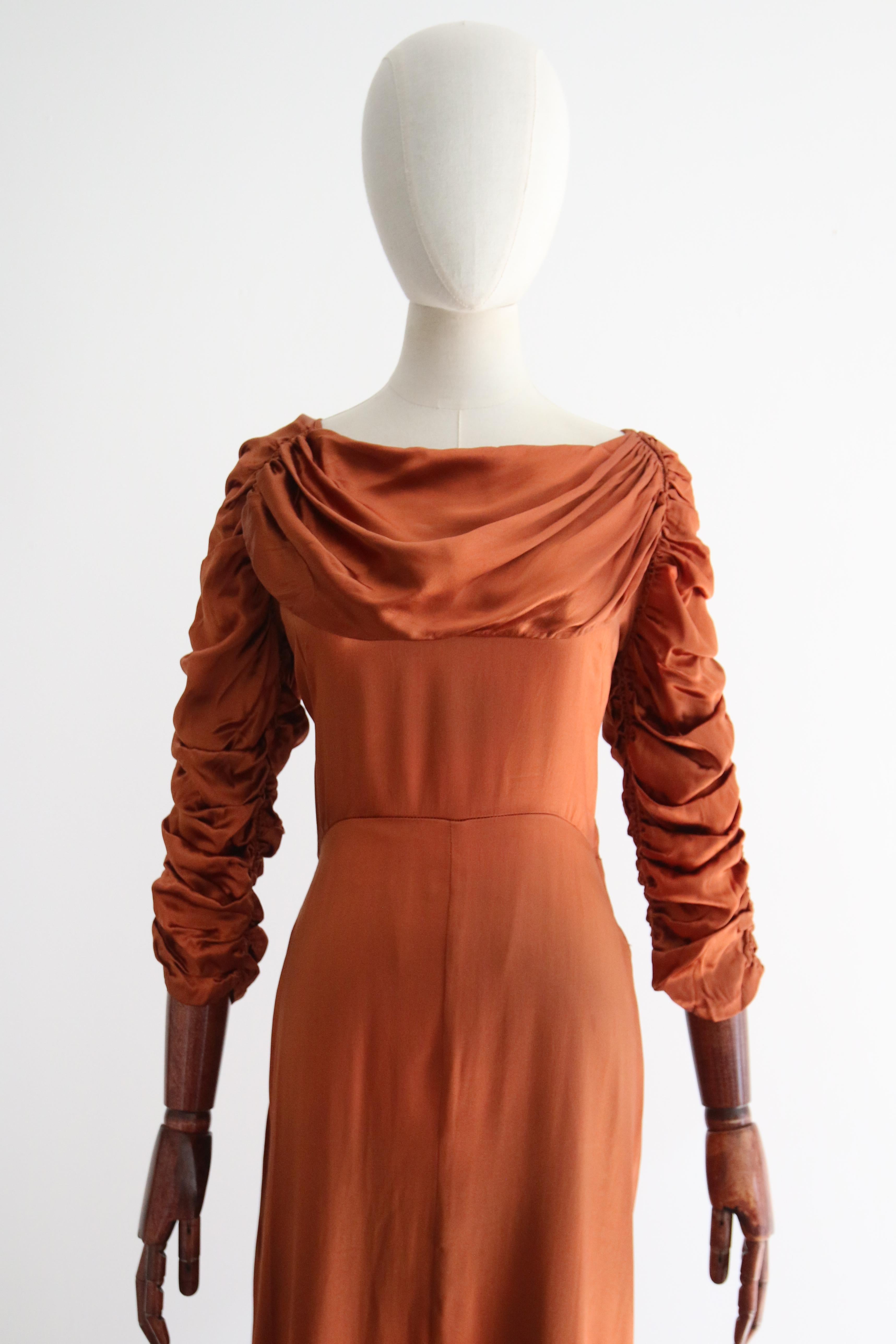 A beauty to behold, this original 1930's satin dress rendered in the most sumptuous shade of amber satin, is the perfect statement piece for your special occasion wardrobe. 

The rounded neckline is framed by a feature cowled panel, which drapes and