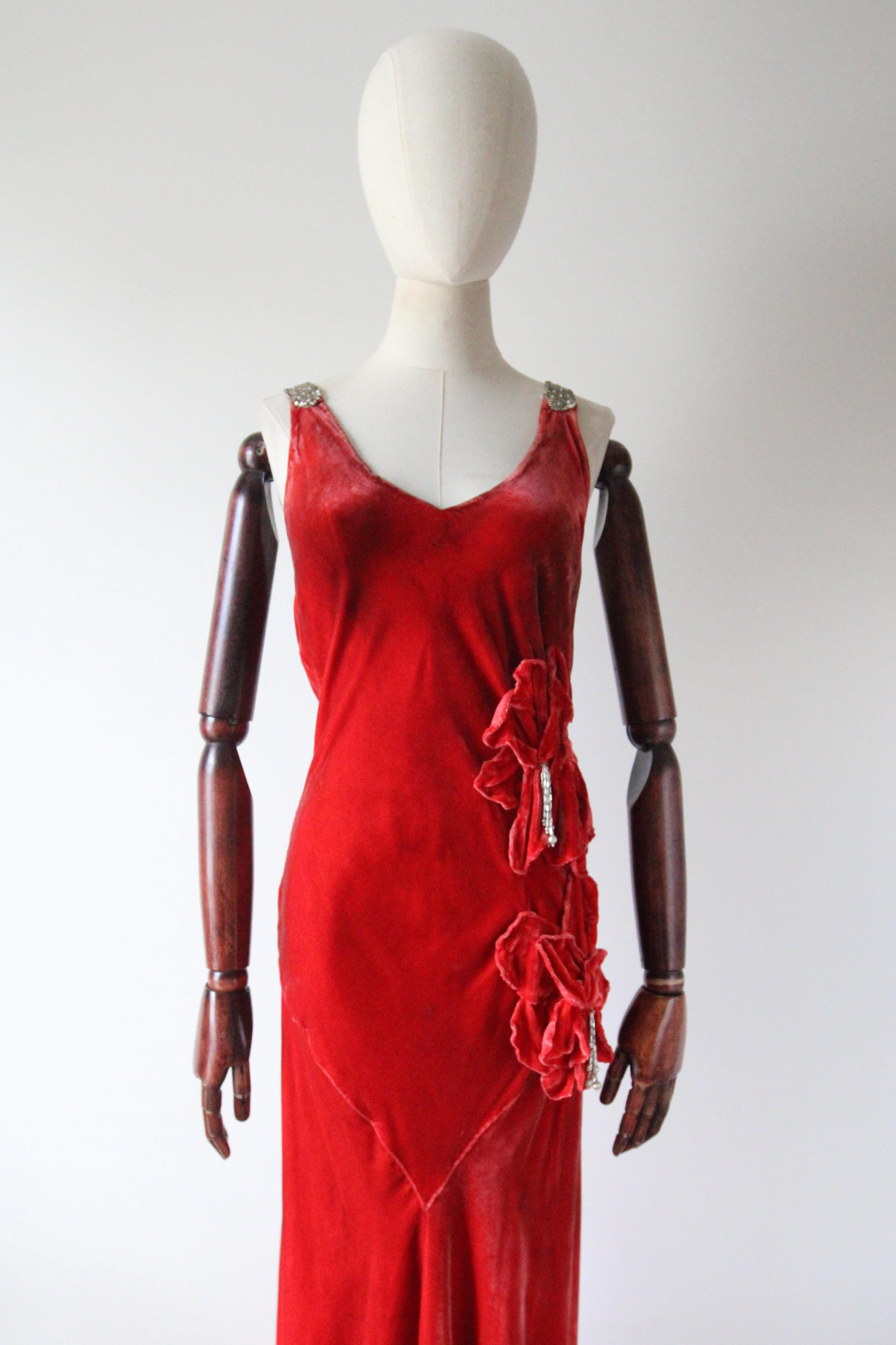 This breathtaking 1930's red silk velvet bias cut dress, decorated with trailing silk velvet hibiscus flowers and rhinestone and glass beadwork, is a rare piece to behold. 
The V shaped neckline, is framed by silk chiffon shoulder straps, which are