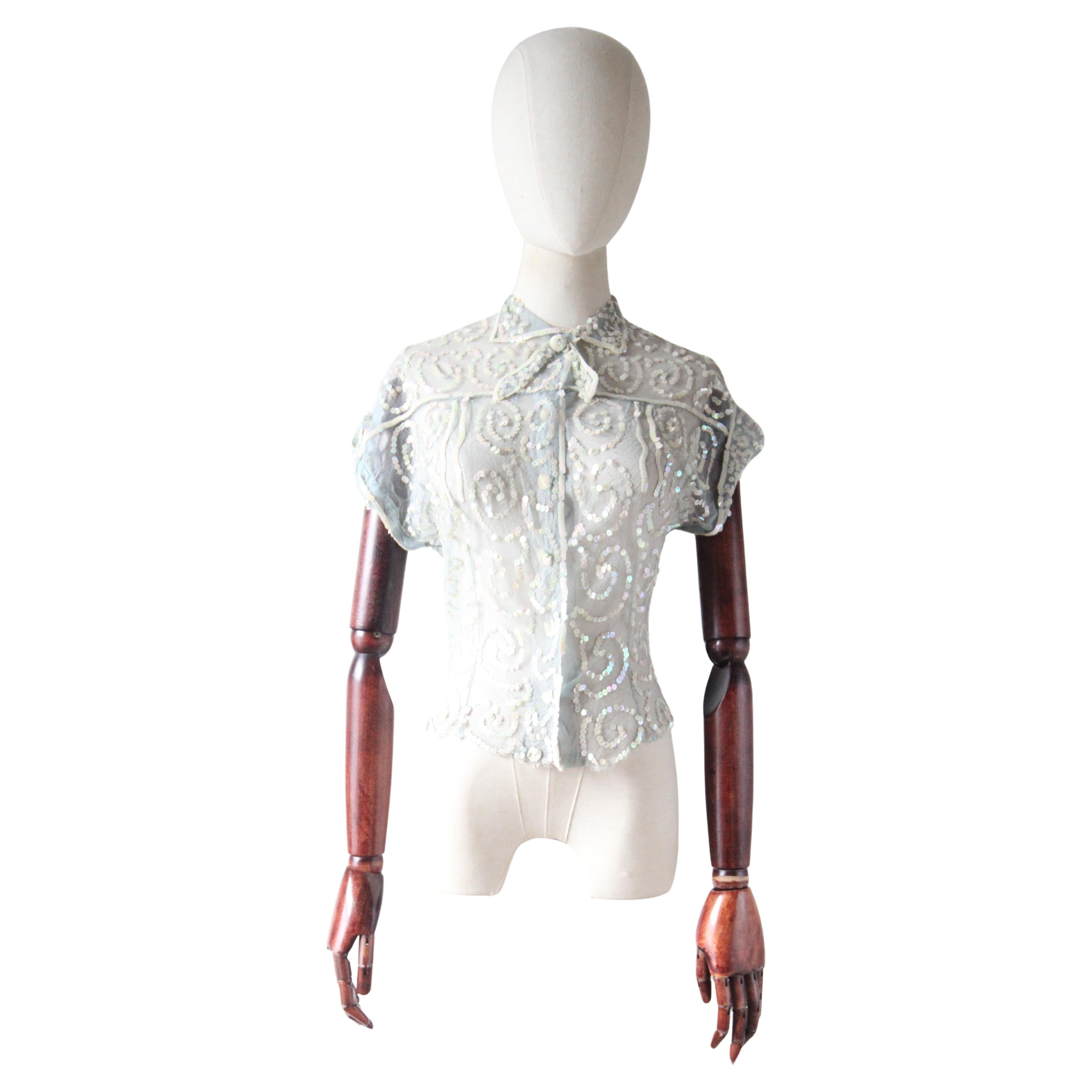 This sparkling 1930's ice blue cotton based tulle blouse, embellished with a swirling pattern of iridescent cream coloured sequins,  is just the perfect touch of sparkled to add your outfit.  

The rounded neckline of the blouse is framed by a