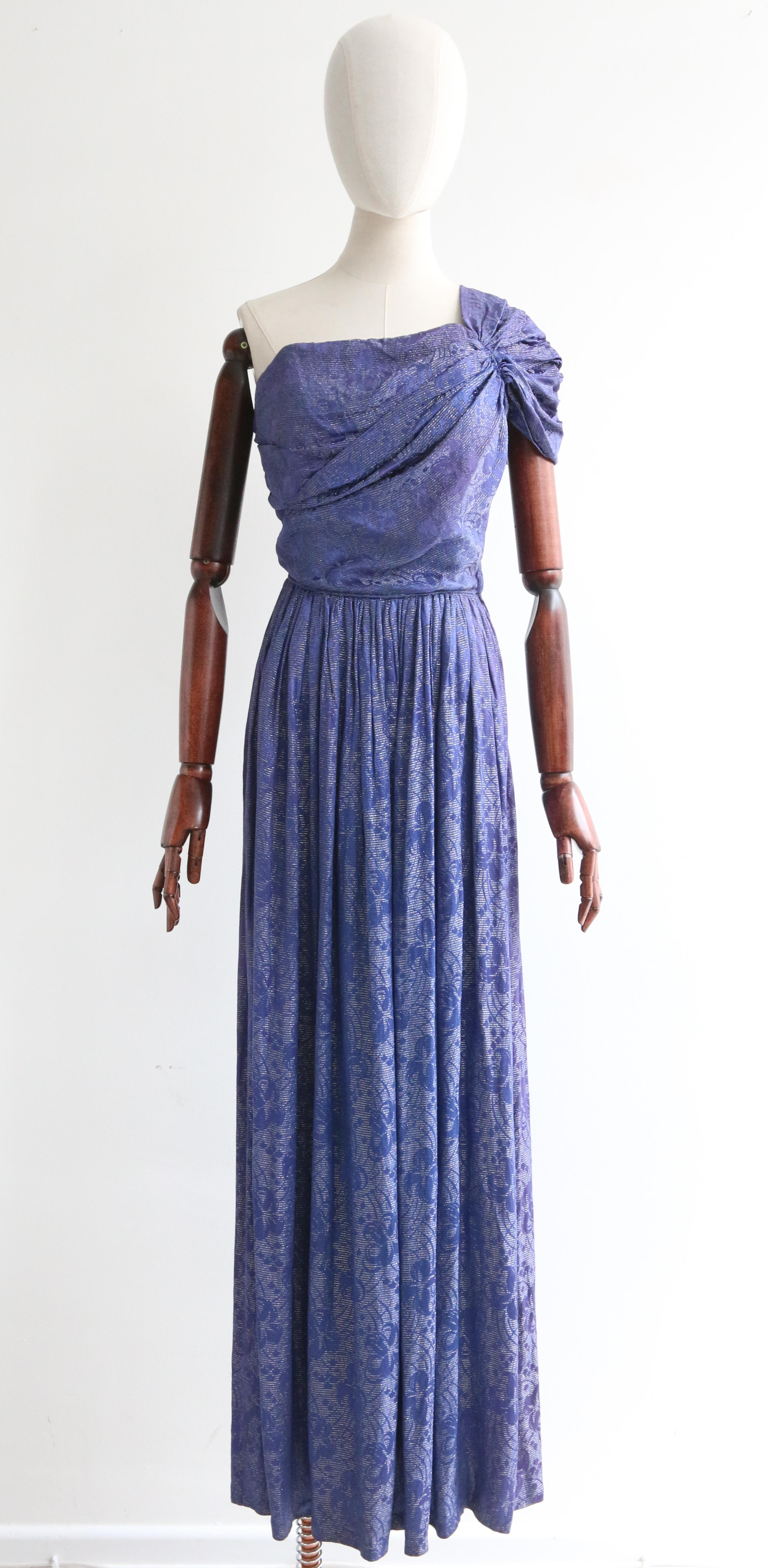 Cette fascinante robe en brocart de soie bleu nuit des années 1930, avec un accent en fil lamé argenté, est une pièce rare à voir

L'encolure droite de la robe est mise en valeur par une magnifique bretelle unique, élégamment plissée et froncée, qui