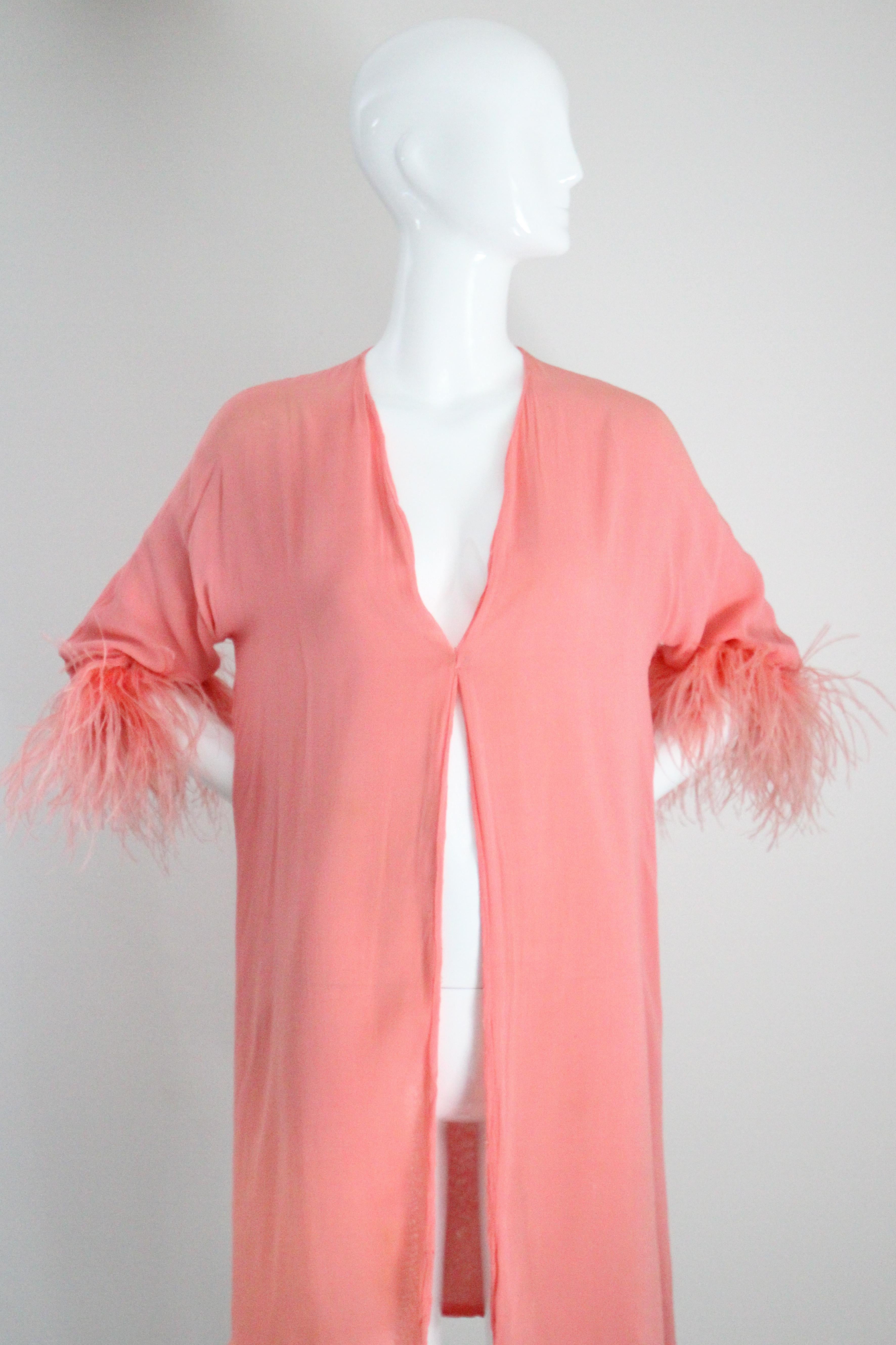 Diese prächtige Originaljacke aus Seidenchiffon aus den 1930er Jahren in einem perfekten Korallenrosa, verziert mit zarten Straußenfederbündchen, ist eine Rarität. 

Der V-förmige Ausschnitt wird mit einem einfachen, verdeckten Haken und Ösen
