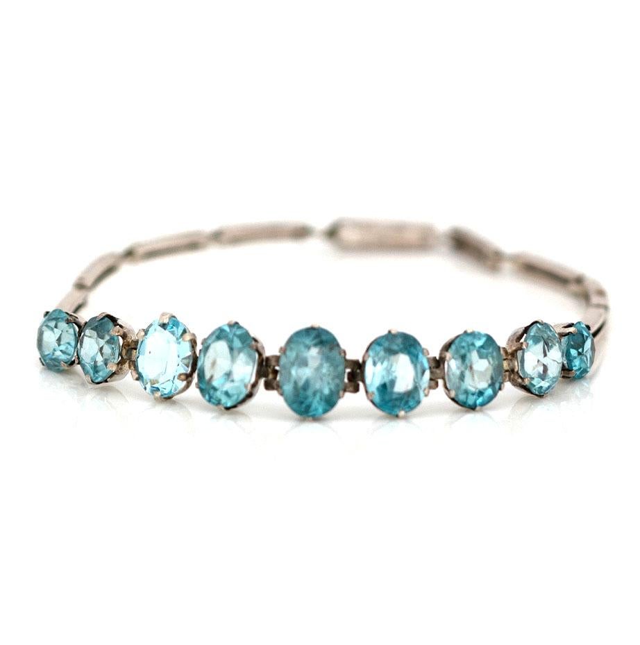Adorn your wrist with this stunning bracelet showcasing nine luminous blue Zircon gemstones, each skillfully cut into round and oval shapes, and nestled within a bright 9ct white gold setting. Adding to its allure, the bracelet fastens securely with