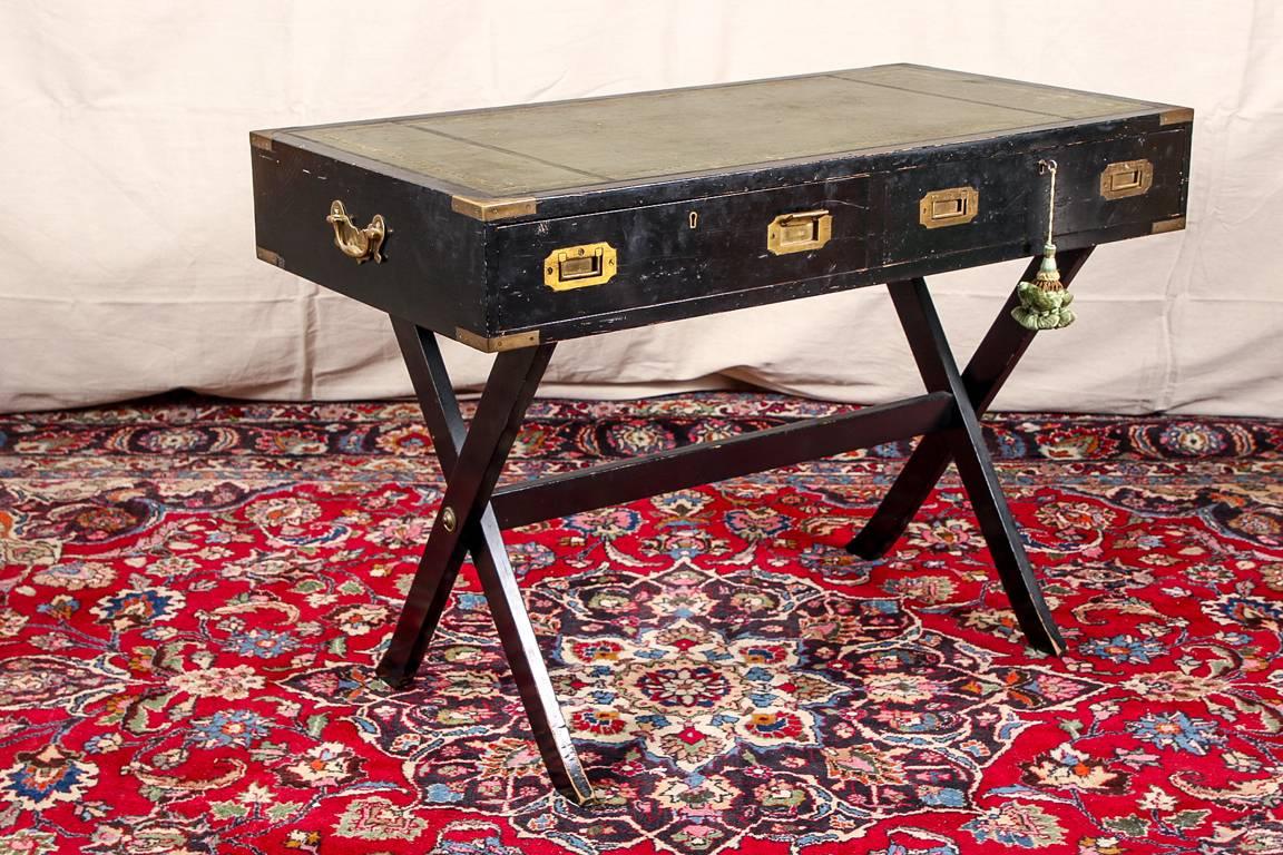 Vintage 1940s ebonized Baker Campaign desk, two apron drawers with brass escutcheons (with a key) and a tooled green leather top, raised on a collapsible ebonized X-frame with stretcher, brass hardware with carry handles on the sides. 

Condition: