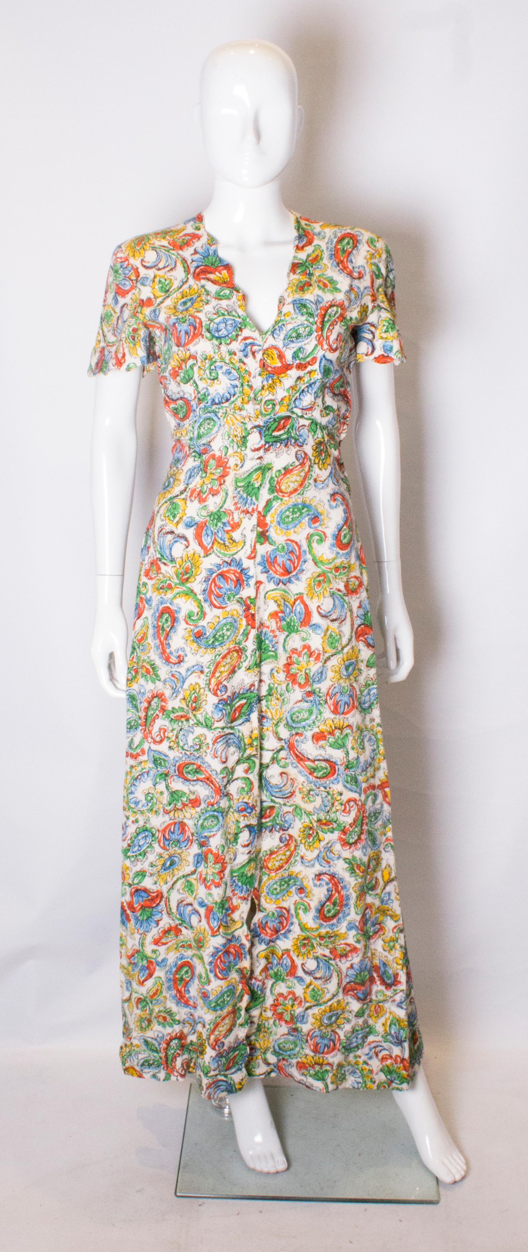 A stunning linen print gown from the 1940s .
The dress has a scallop edged neckline and sleaves with a popper opening at the side. it has a white background with multi colour print.