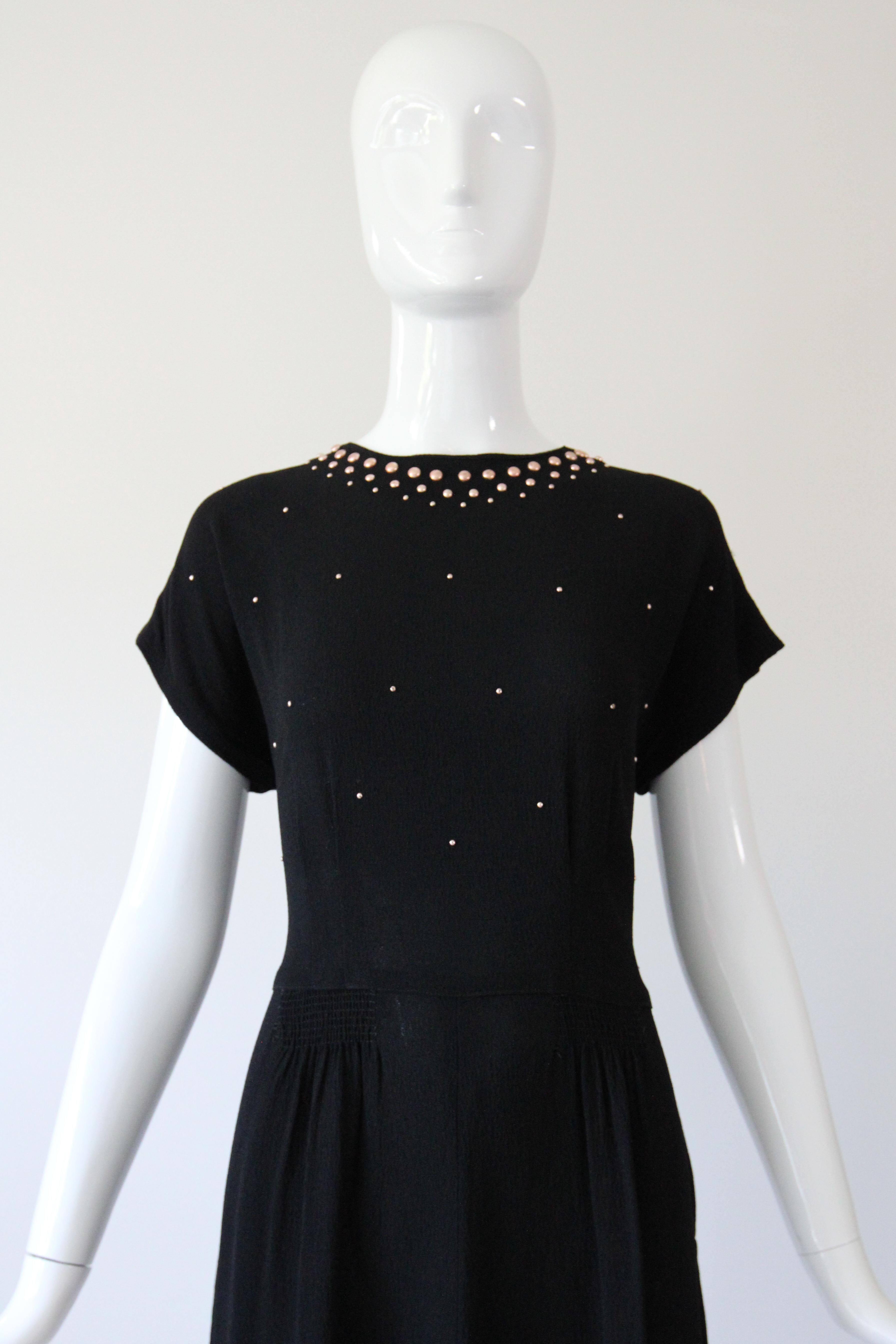 Dieses originelle schwarze Seidenkleid aus Crêpe aus den 1940er Jahren in einer wahrhaft ikonischen Silhouette ist mit roségoldfarbenen Metallnieten verziert und genau das Richtige für Ihre Cocktailsammlung.  

Der runde Ausschnitt des Kleides wird