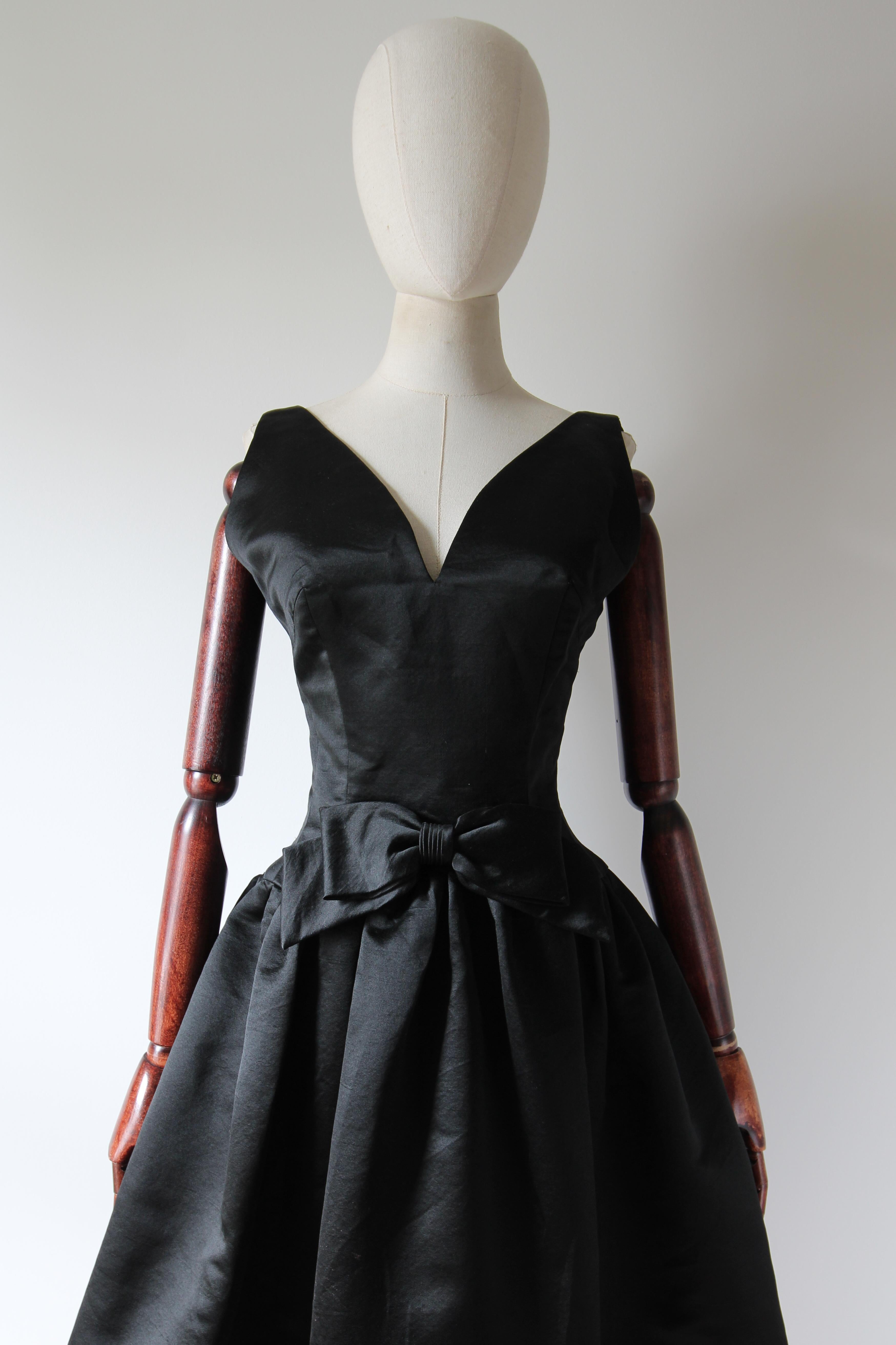 This truly mesmerising 1950's couture dress, rendered in a sumptuous black satin and accented by a decorative central bow, is just the piece to add to your timeless capsule collection.

The elegant curved V shaped neckline is framed by medium