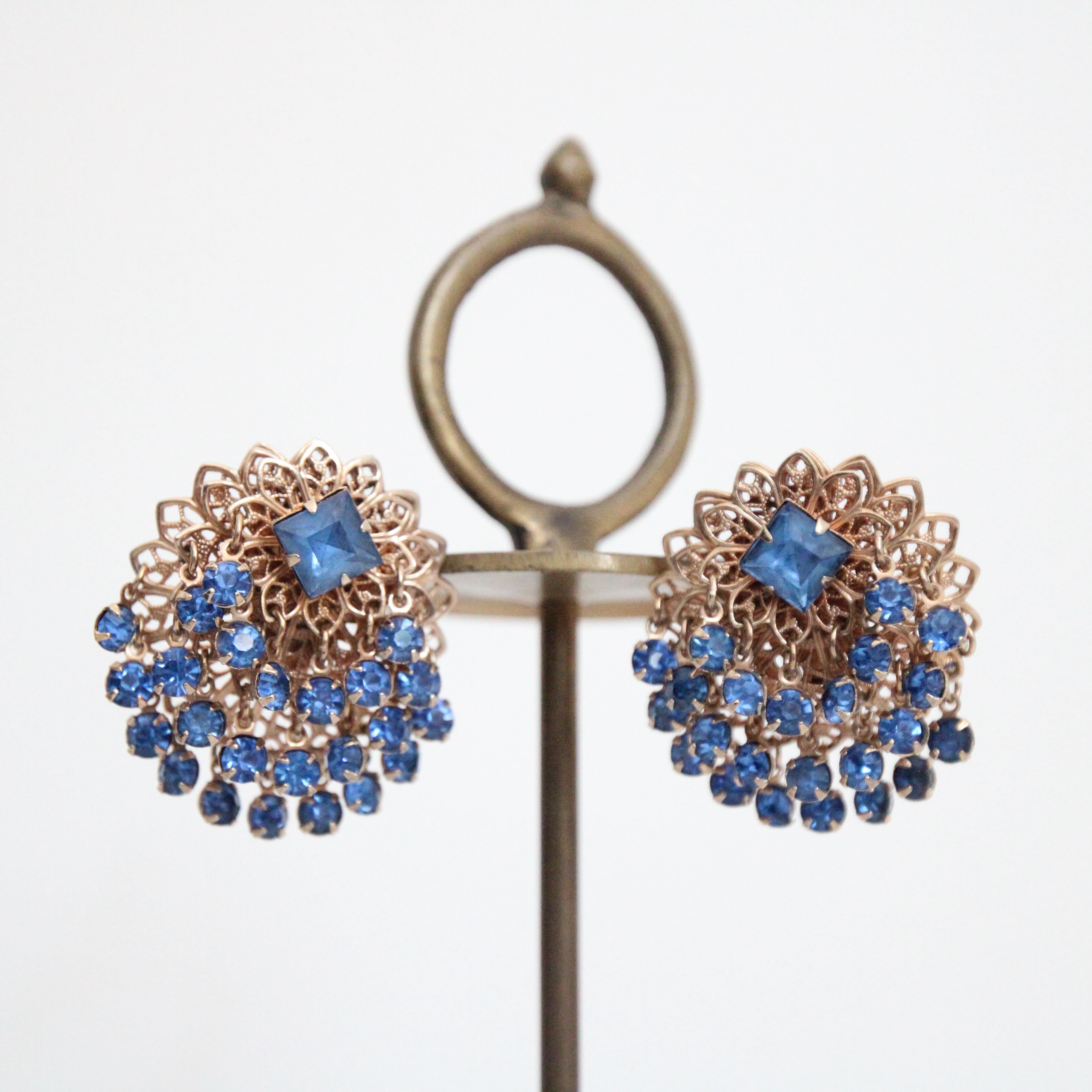 Dieses wirklich auffällige Paar Ohrringe aus den 1950er Jahren aus goldfarbenem Metall mit blauen Strasssteinen im Krallenbesatz ist das perfekte Accessoire für Ihr Outfit. Die goldfarbenen, filigranen Sockel sind mit drei aufeinander abgestimmten,