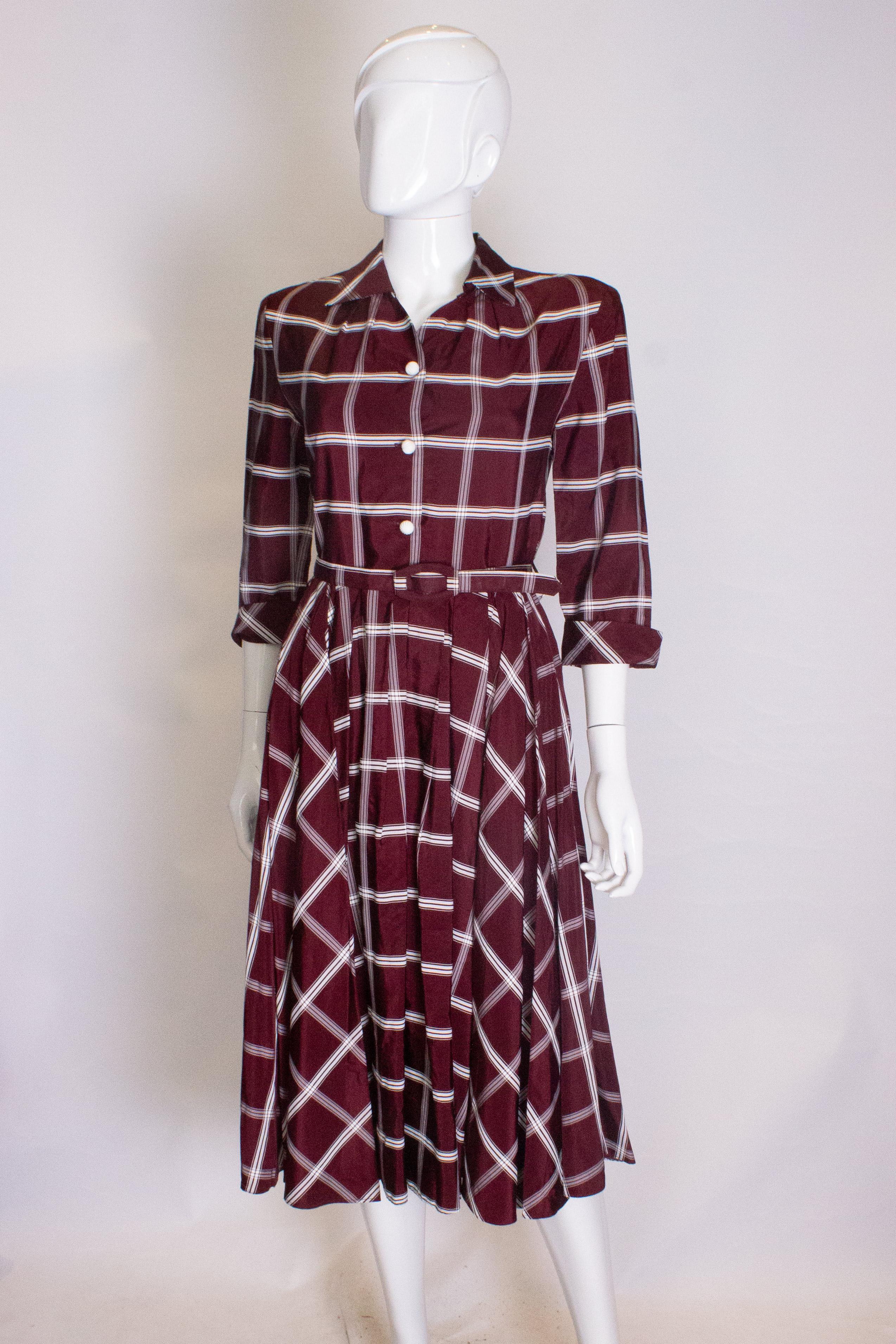 A head turning shirt dress in burgundy with white detail. The dress has a shirt collar, v neckline and button front opening. It has elbow length sleeves with turn back cuffs, self fabric belt and pleats at the waist.