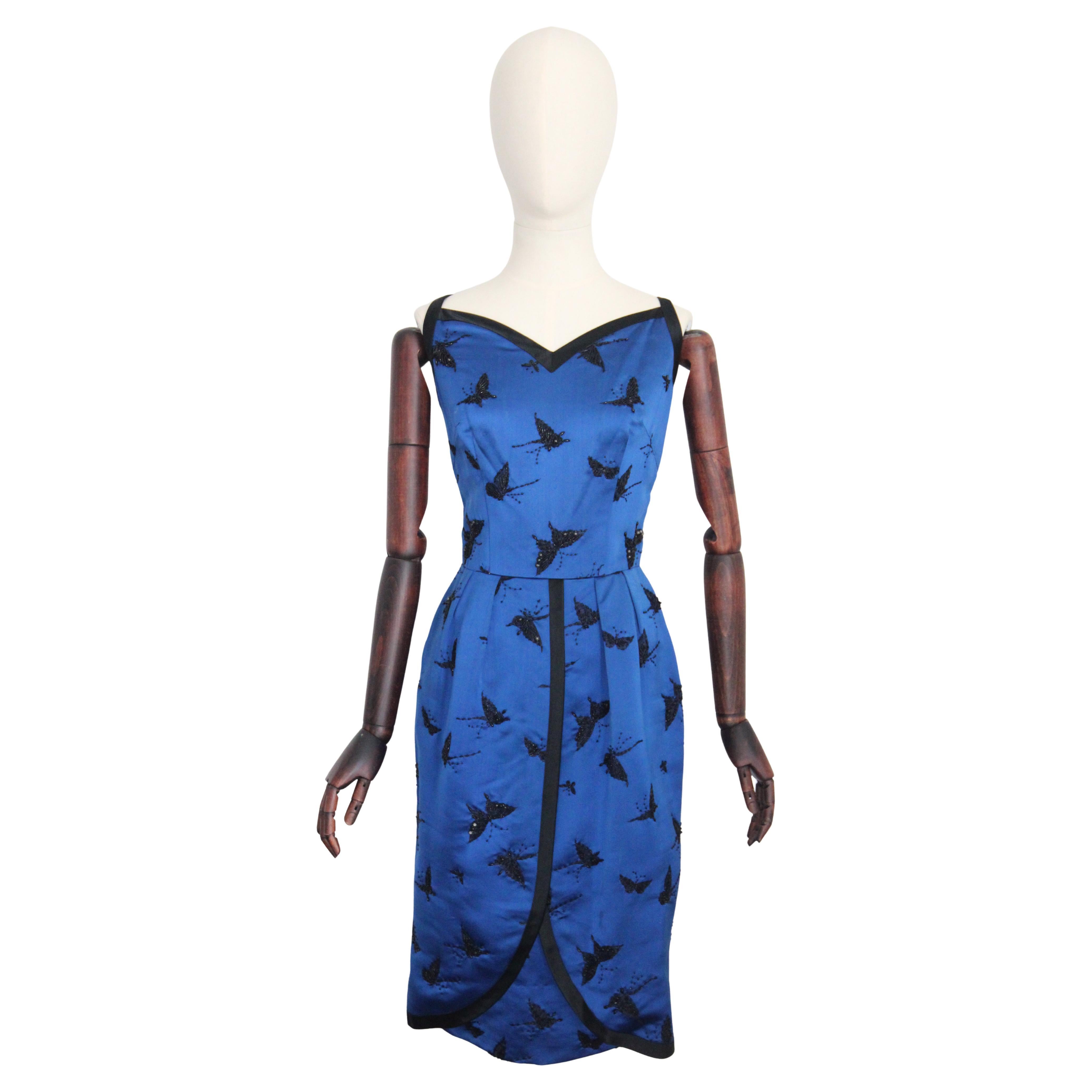 Este espectacular vestido de cóctel de los años 50, confeccionado en el tono más llamativo de satén azul cobalto, con detalles de satén negro en contraste y un delicado trabajo de pedrería negra en forma de mariposas revoloteando sobre motivos de