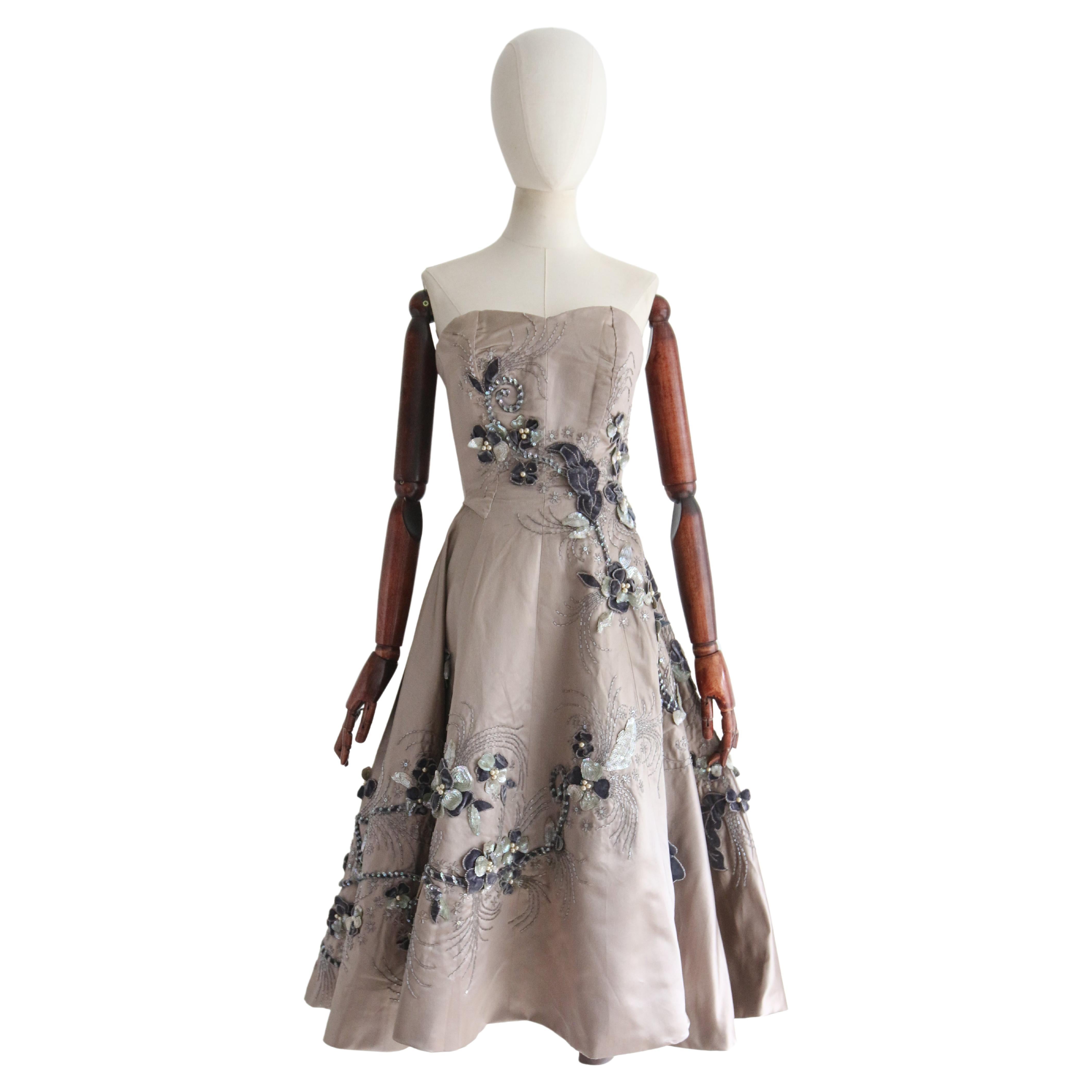 Rendered in the most exquisite stone coloured satin and embellished with silver glass bugle beads, pearlescent beads, iridescent sequins, and silk velvet, depicting a wild and blooming trailing floral pattern, this original 1950's couture dress is a