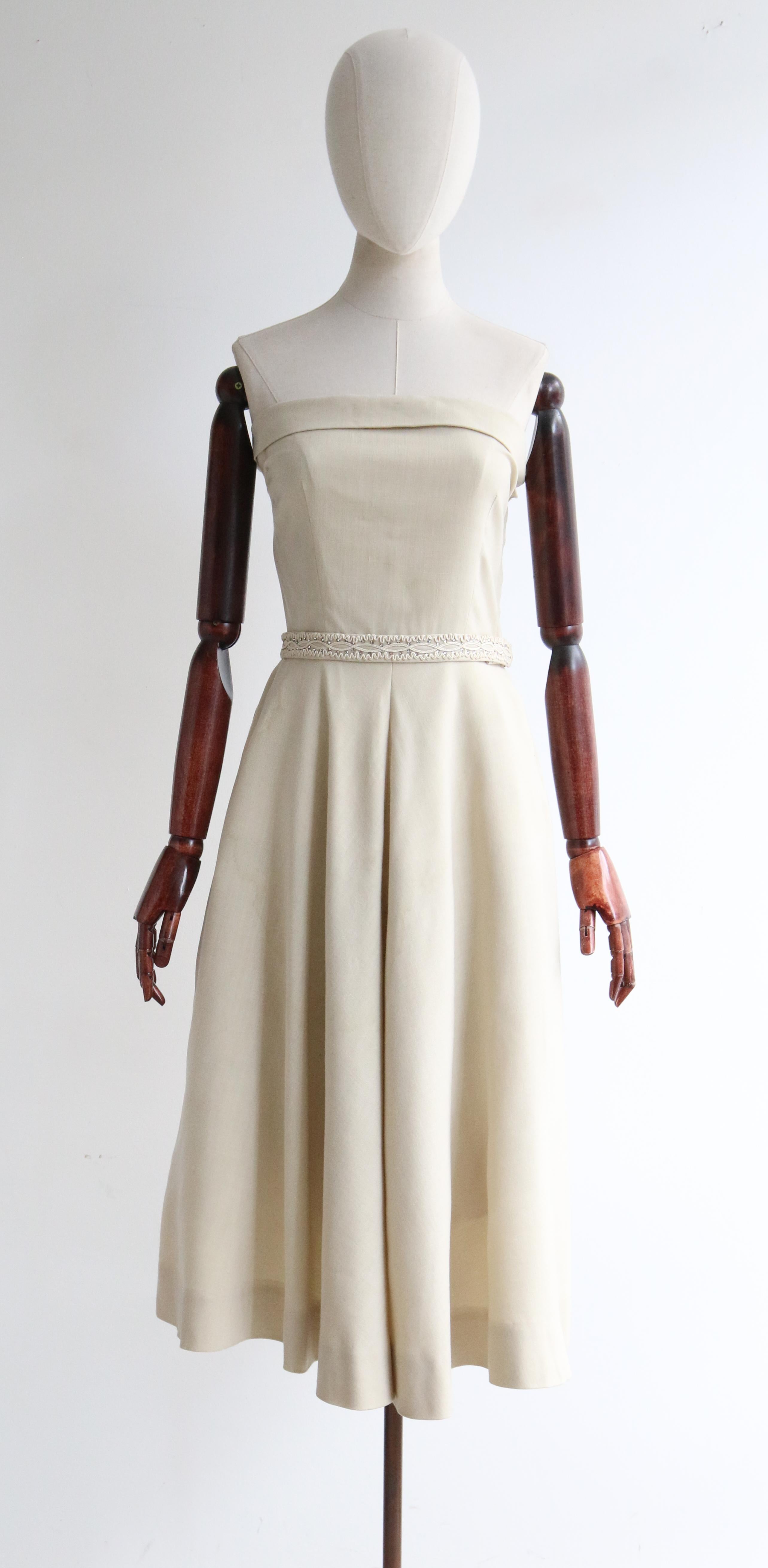 Este magnífico vestido crema de los años 50 con bolero a juego, adornado con soutache y brillantes plateados engarzados, es la pieza perfecta para una ocasión especial. 

El corte recto del escote, sin tirantes, se acentúa con un cuello vuelto que