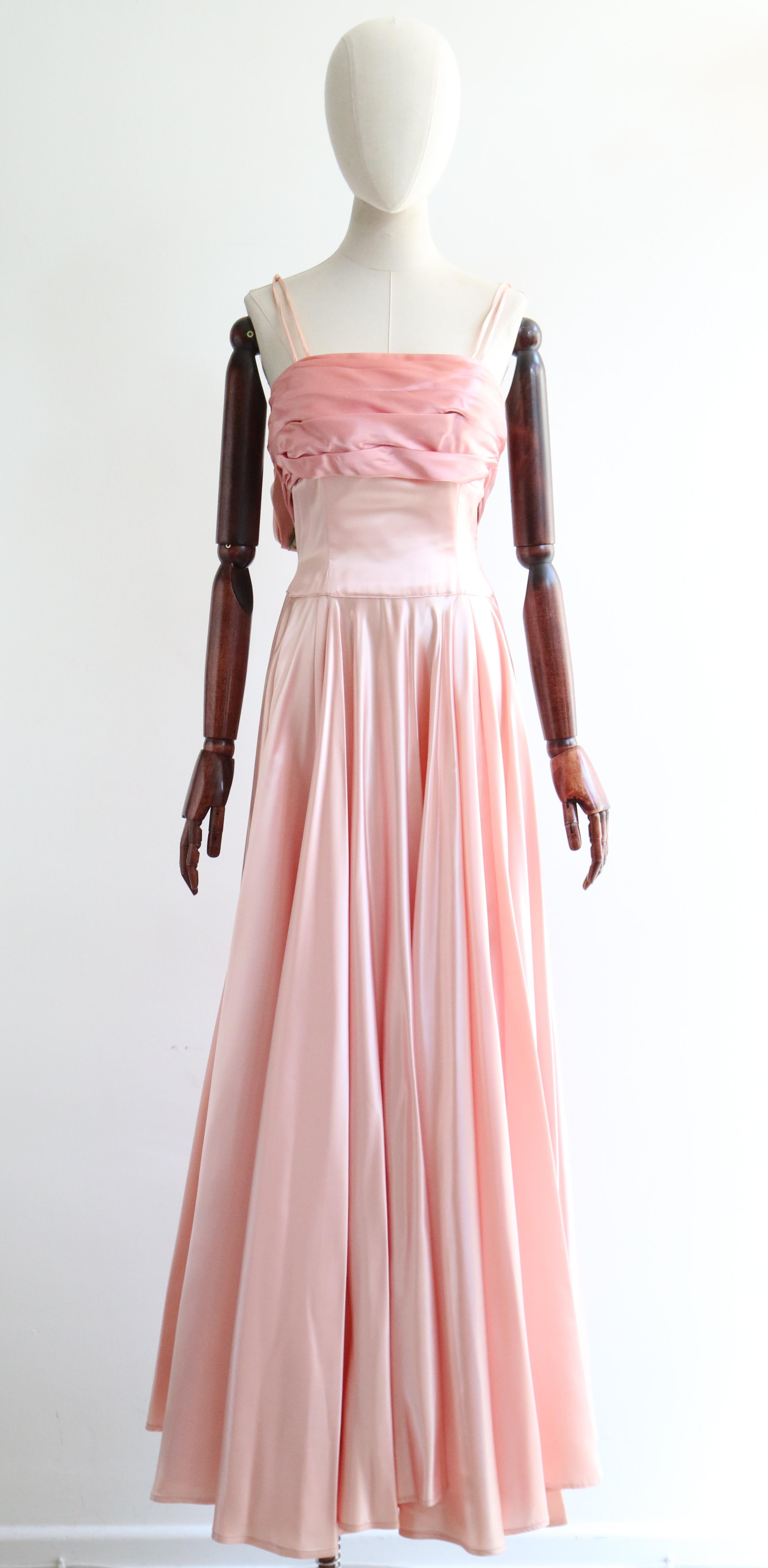 This breathtaking 1950's duchess satin gown, in the most eye-catching shade of petal pink and blush pink, is the perfect piece for that special occasion.  

The straight cut of the neckline is framed by thin double shoulder straps that twist