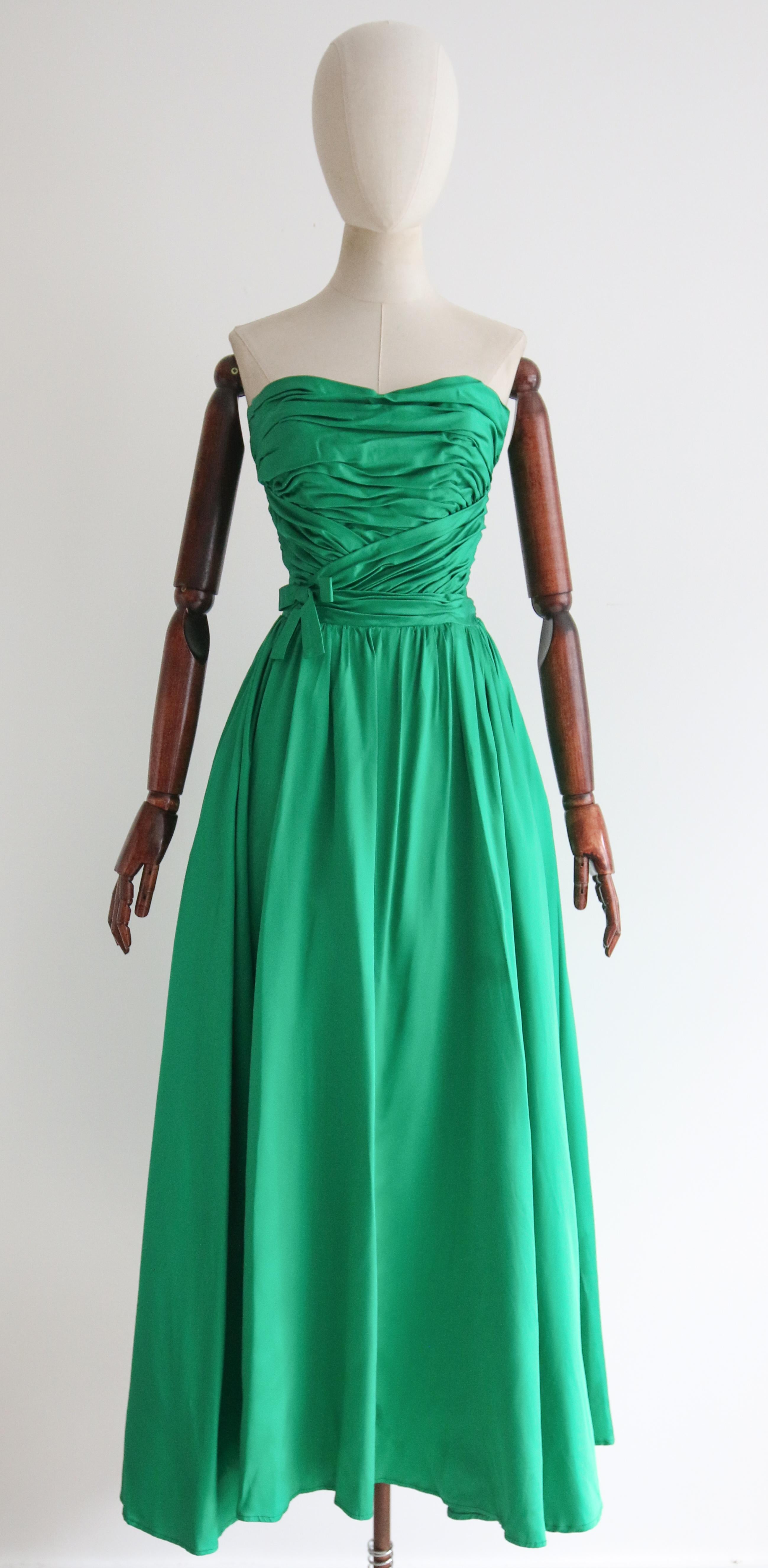 This breathtaking 1950's duchess satin gown, in the most eye-catching shade of emerald green, accented by gathered pleats and a decorative bow, is the perfect piece for that special occasion. 

The strapless neckline of the dress, is set off by a