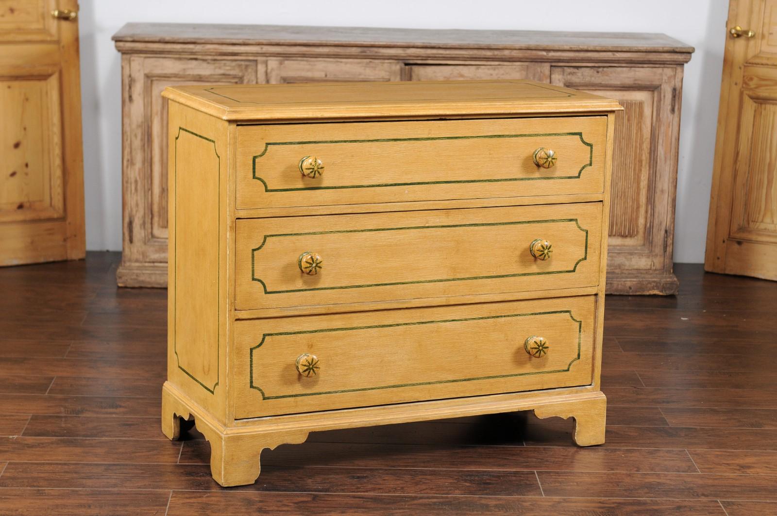 An English vintage painted three-drawer commode from the mid-20th century, with muted yellow finish and painted cartouches. Born in England during the midcentury period, this charming painted commode features a rectangular top sitting above three