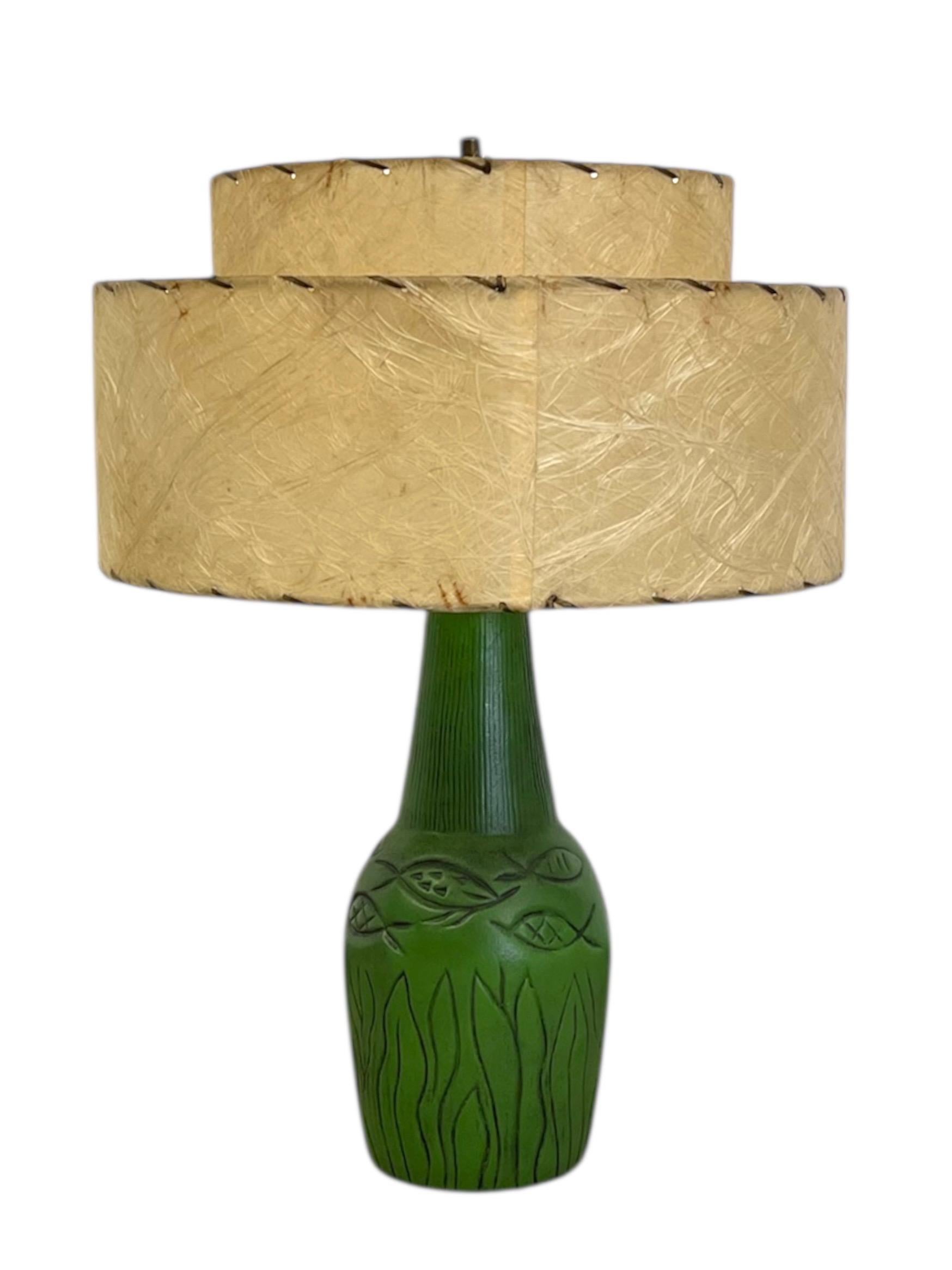 Lampada da tavolo di metà secolo in materiale simile alla ceramica, con una base verde brillante con motivi di pesci e piante acquatiche intagliati a mano. La lampada conserva il paralume originale in fibra di vetro a due livelli, con bordi a