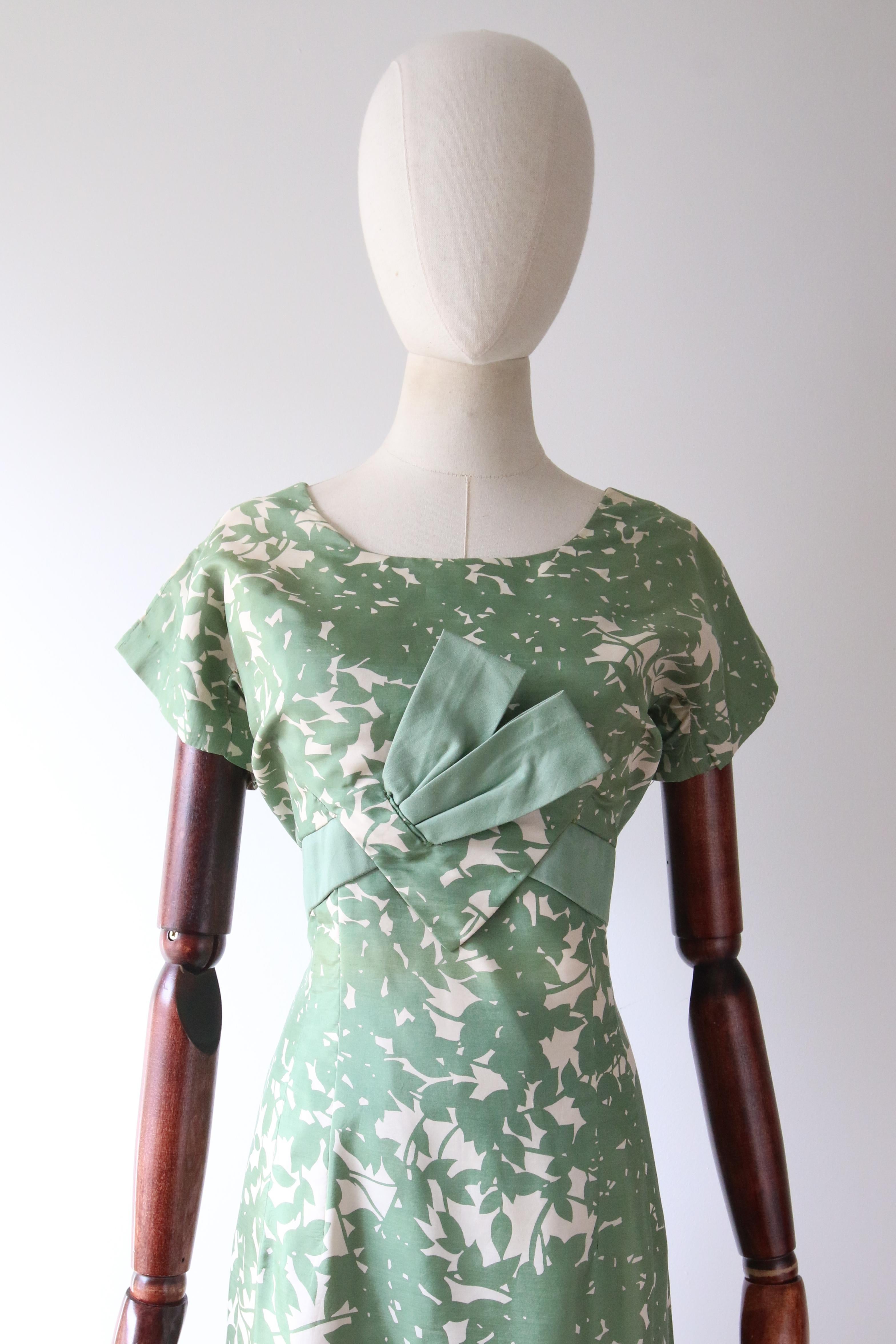 This enchanting 1950's green and cream silk dress in an abstract pattern of leaves, set off by a decorative bow panel along the bodice,  is the perfect piece to welcome the new season with.

The rounded neckline is framed by a simple shoulder line