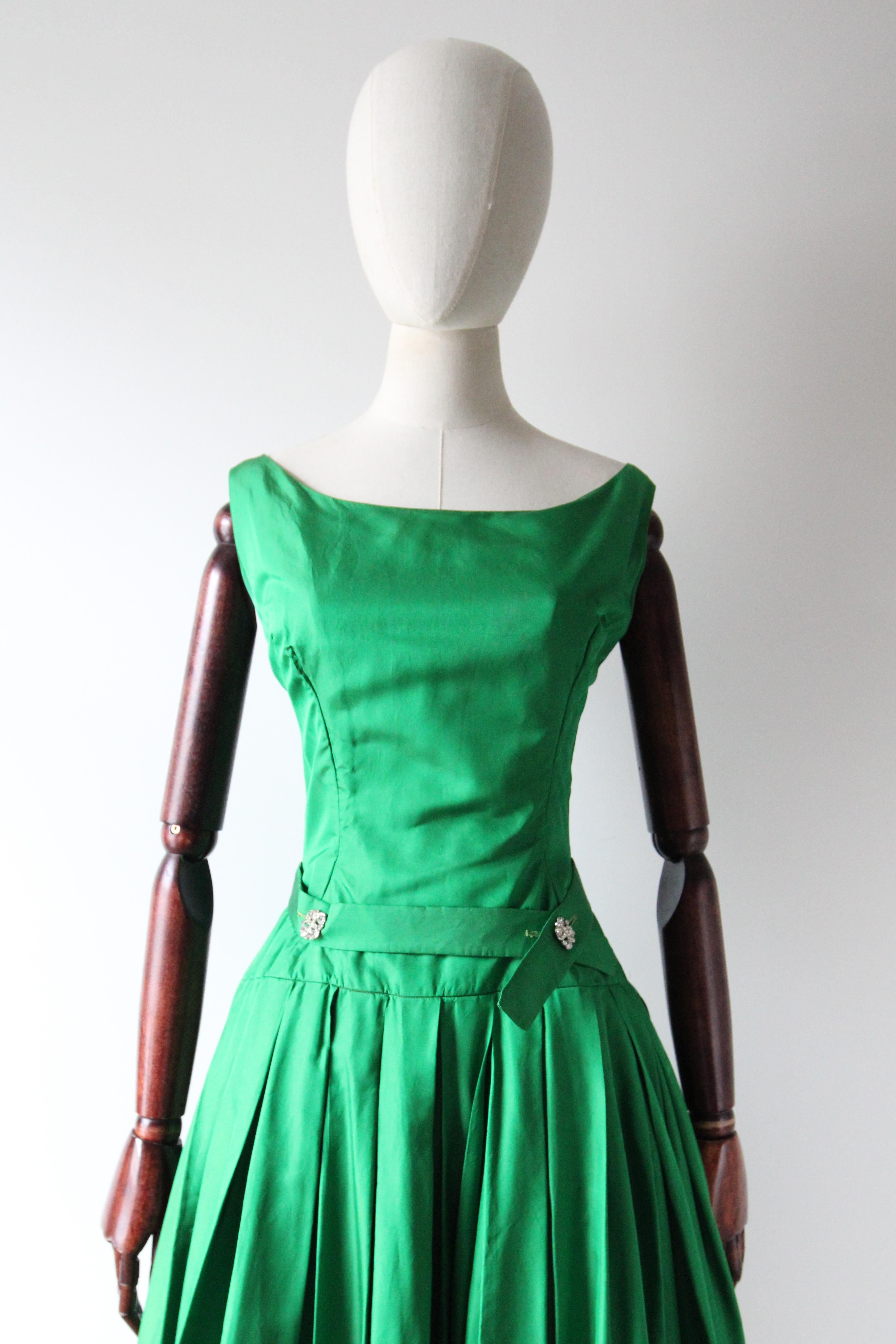 Dieses bezaubernde Kleid von Suzy Perette aus den 1950er Jahren in einem leuchtenden, immergrünen Seidenton mit silbernen Strassknöpfen ist genau das richtige Stück für Ihre Vintage-Sammlung. 

Der runde Bateau-Ausschnitt wird von schlichten