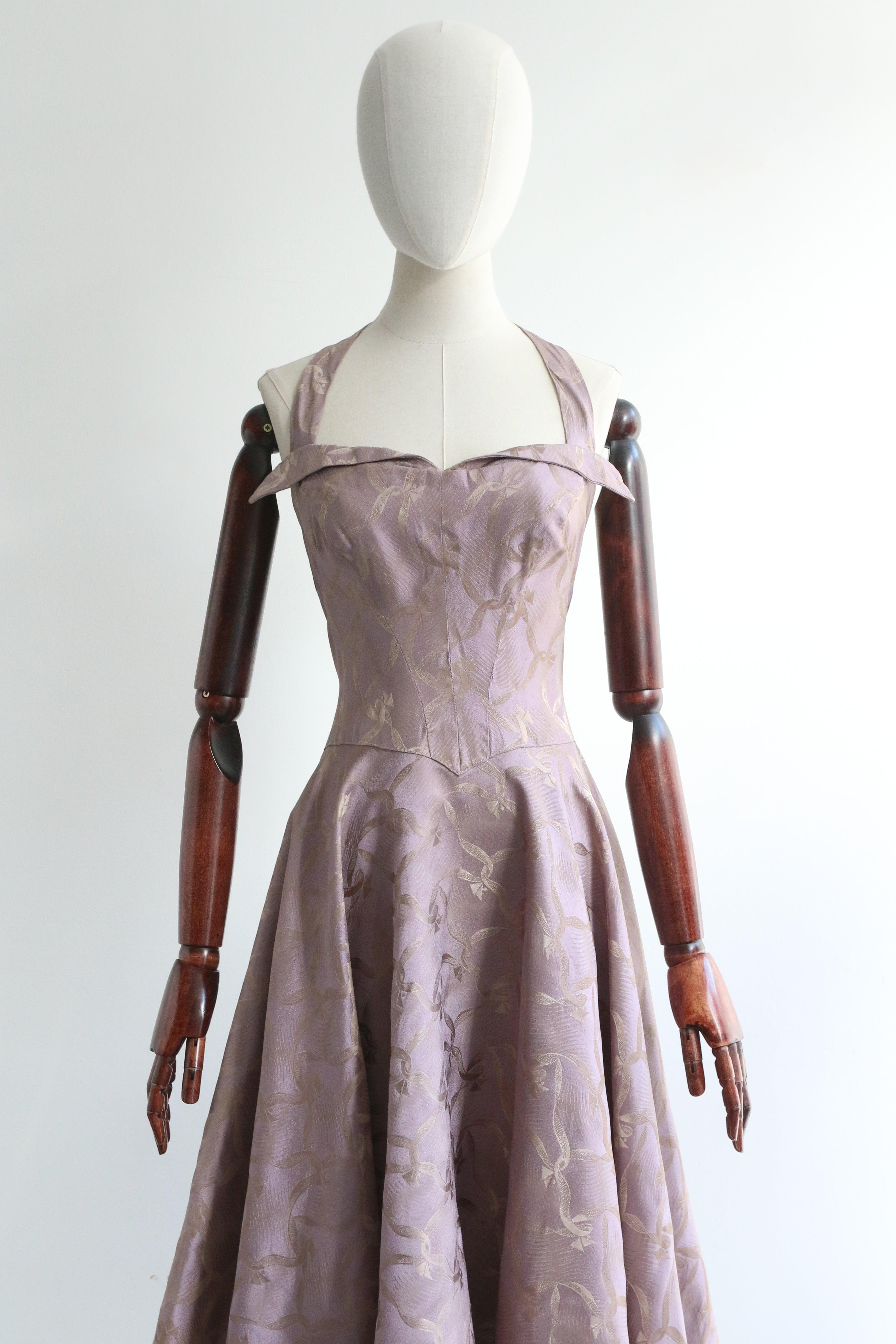 Confeccionado en los tonos lila y gris más suntuosos con un diseño de brocado de seda de cintas entrelazadas, este original vestido de cuello halter de los años 50 es la pieza perfecta para tu vestuario de cóctel.

El escote halter enmarca un escote