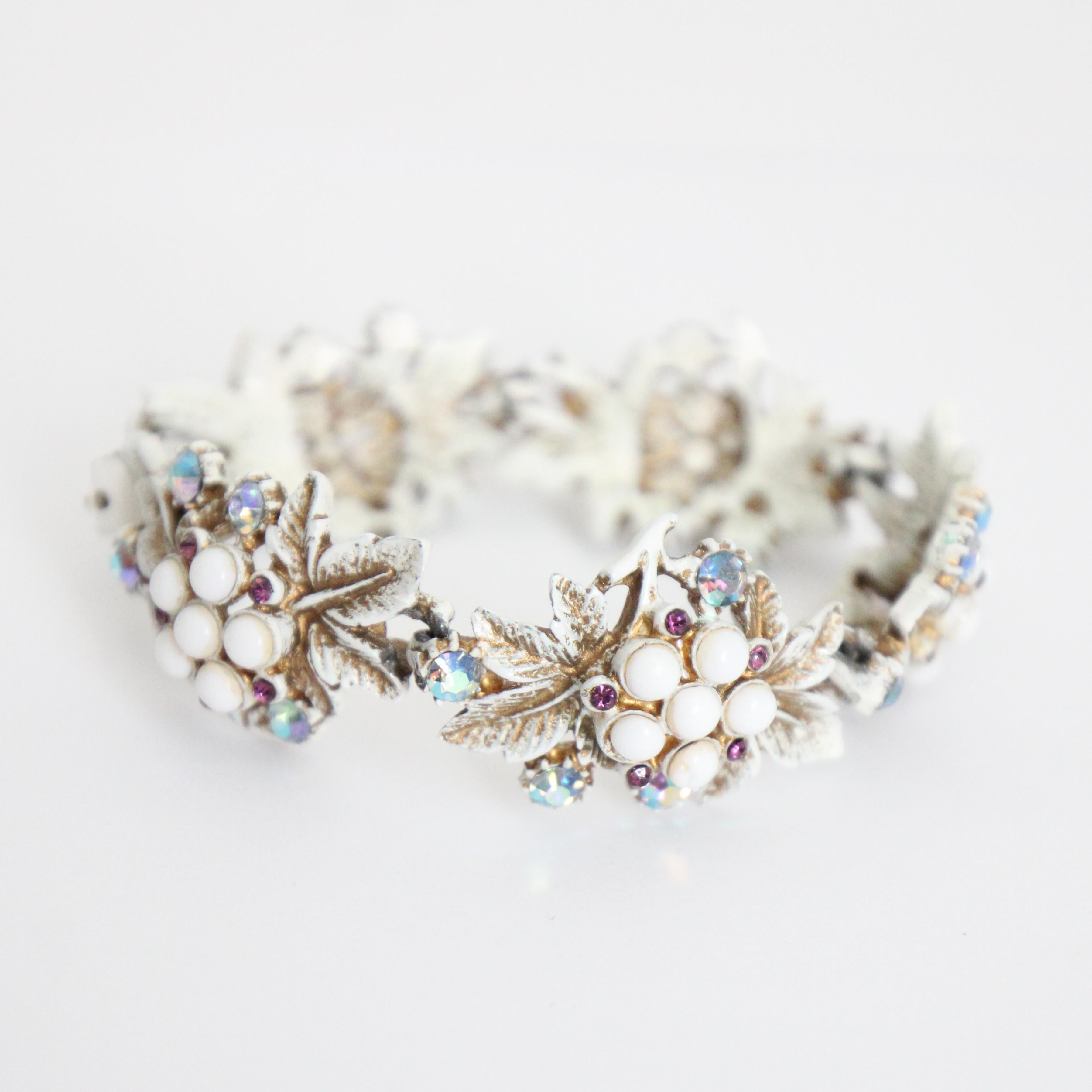 Ce magnifique bracelet floral des années 1950, construit selon un modèle à maillons, qui présente des motifs de feuilles et de fleurs en émail blanc et or, accentués par des cabochons centraux en verre de lait entourés de petits strass violets