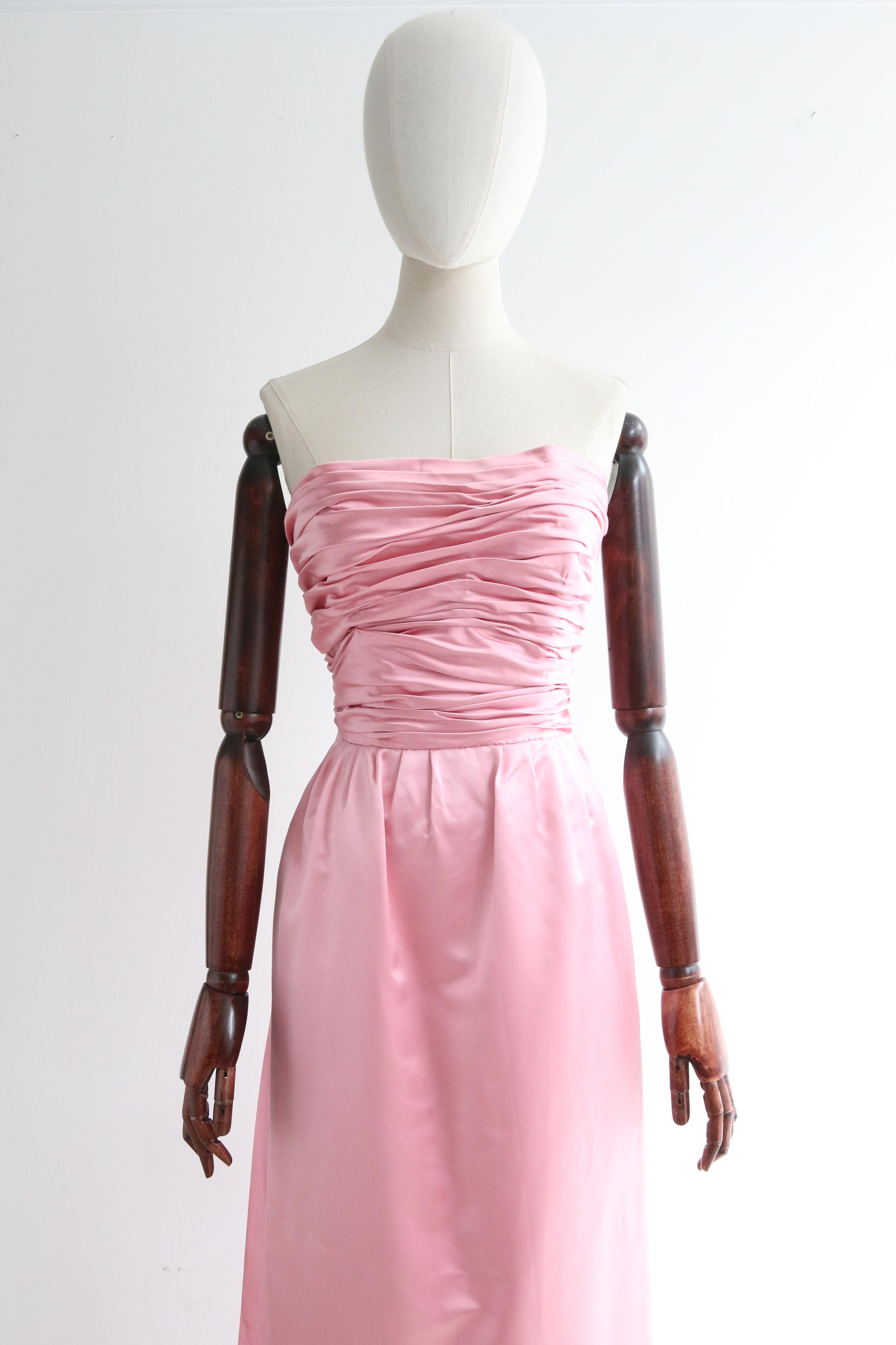 This breathtaking 1950's duchess satin gown, in the most eye-catching shade of petal pink, is the perfect piece for that special occasion. 

The strapless neckline of the dress, is set off by a fitted and boned bodice which boasts elegant gathered