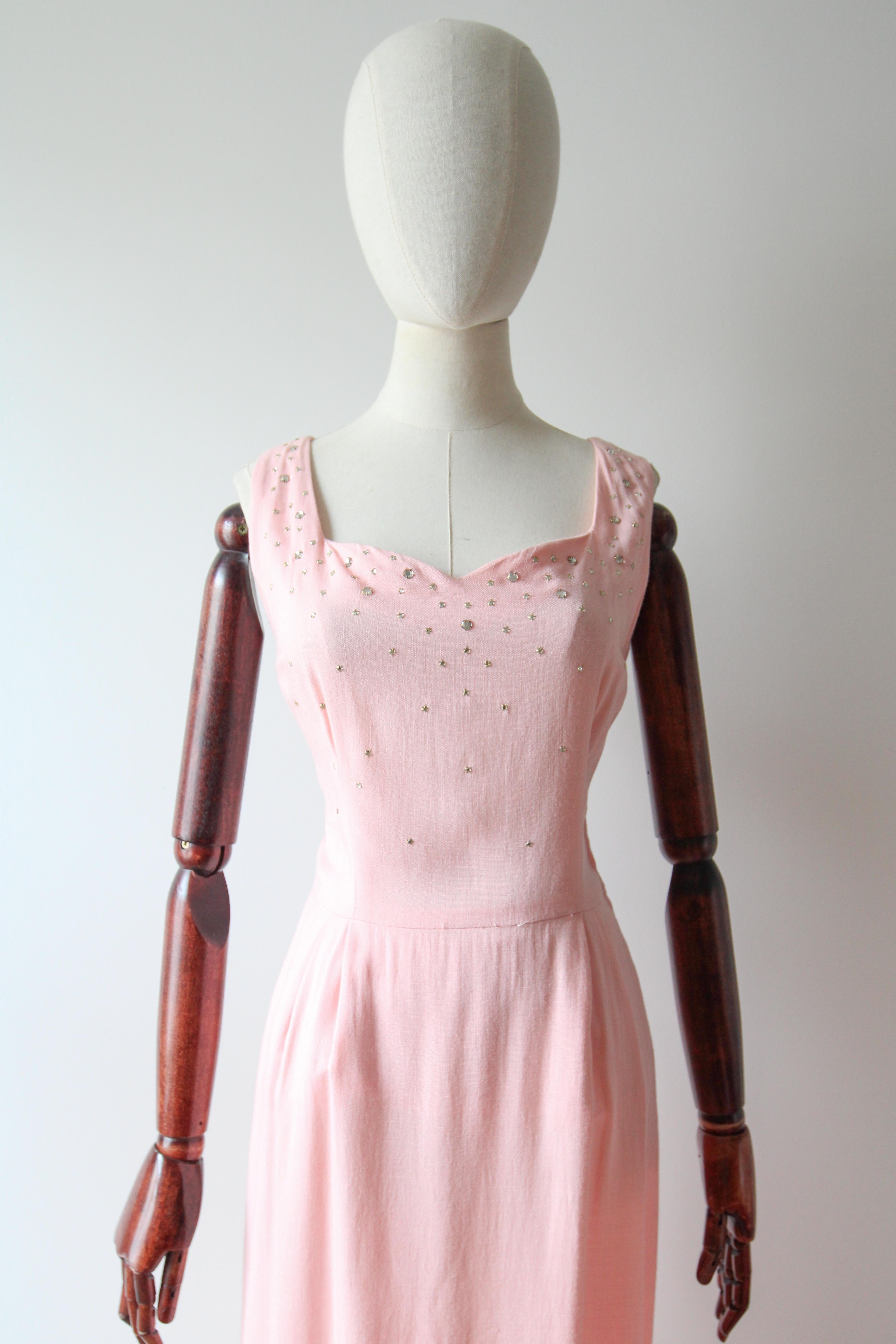 Confeccionado en el tono más suave de algodón rosa y adornado con brillantes de cristal plateados, este vestido de los años 50 es la pieza perfecta para añadir a tu colección vintage.

El escote en forma de corazón está enmarcado por tirantes de