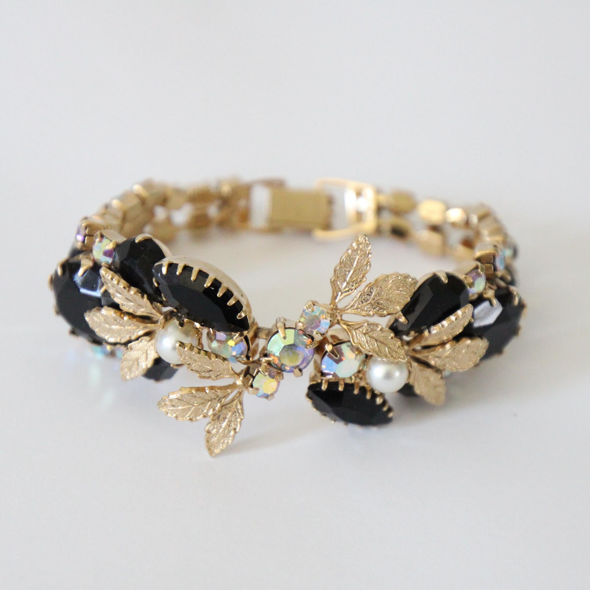 Questo spettacolare braccialetto anni '50, su base metallica dorata, è un pezzo scintillante da aggiungere alla tua collezione di gioielli. 

Il design centrale a foglie strascicate in metallo dorato spazzolato è impreziosito da strass iridescenti