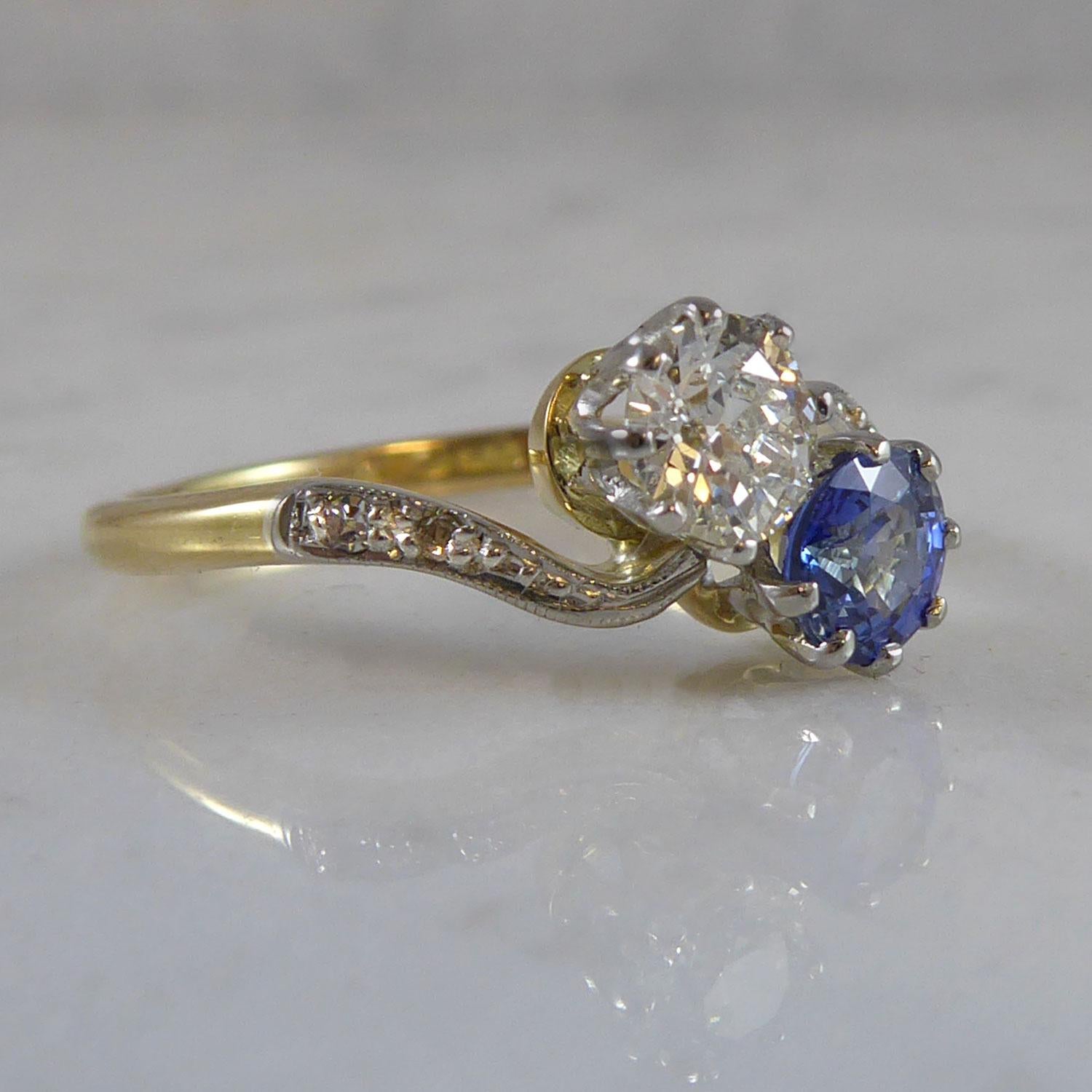 A mid-20th century diamond and sapphire two stone cross over ring.  Set with an old European cut diamond and a round mixed cut sapphire within white basket settings to tapering twist shoulders each set with two single cut diamonds.  Plain polished
