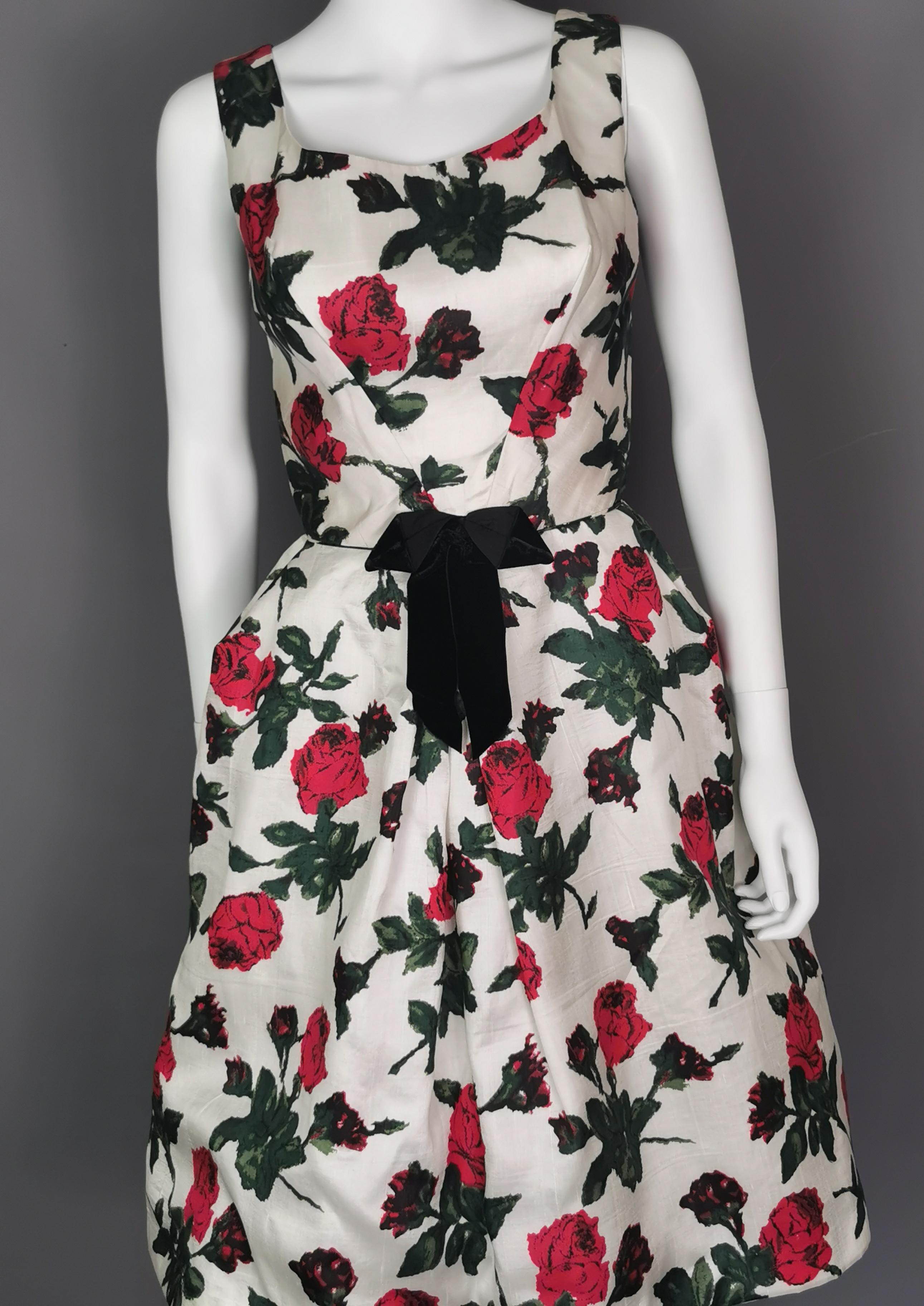 A gorgeous vintage c1950s floral dress.

It is made from silk with a white ground and has an all over Rose print in red and green with black accents.

The dress is fully lined and has a full skirt and fitted bodice, it is sleeveless and has a small
