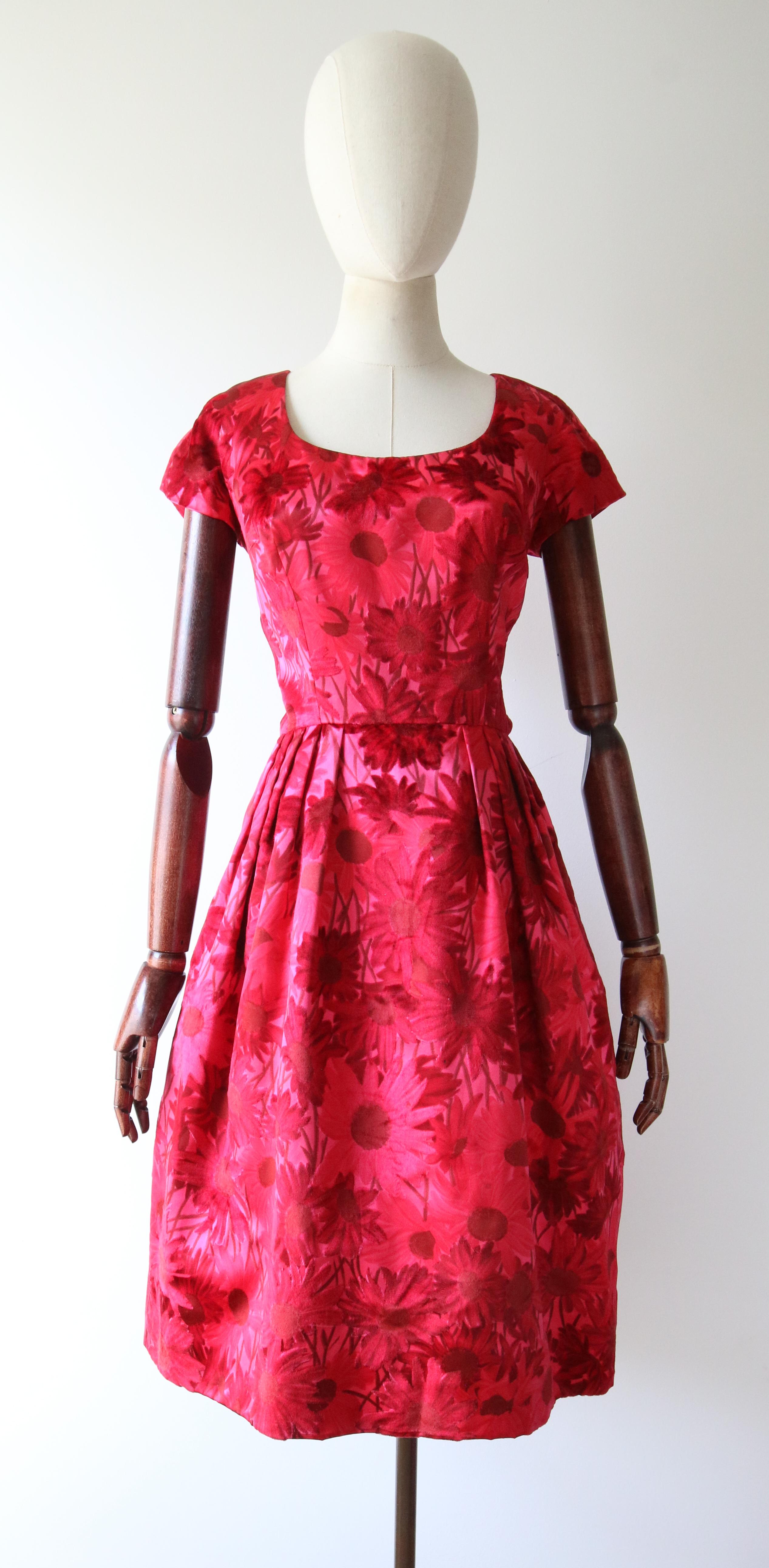 A gem to behold, this original 1950's satin dress, in shades of pink and red accented by silk velvet flocking details, in a pattern of wild daisies, is just the perfect piece for your vintage collection.

The rounded neckline of the dress is framed