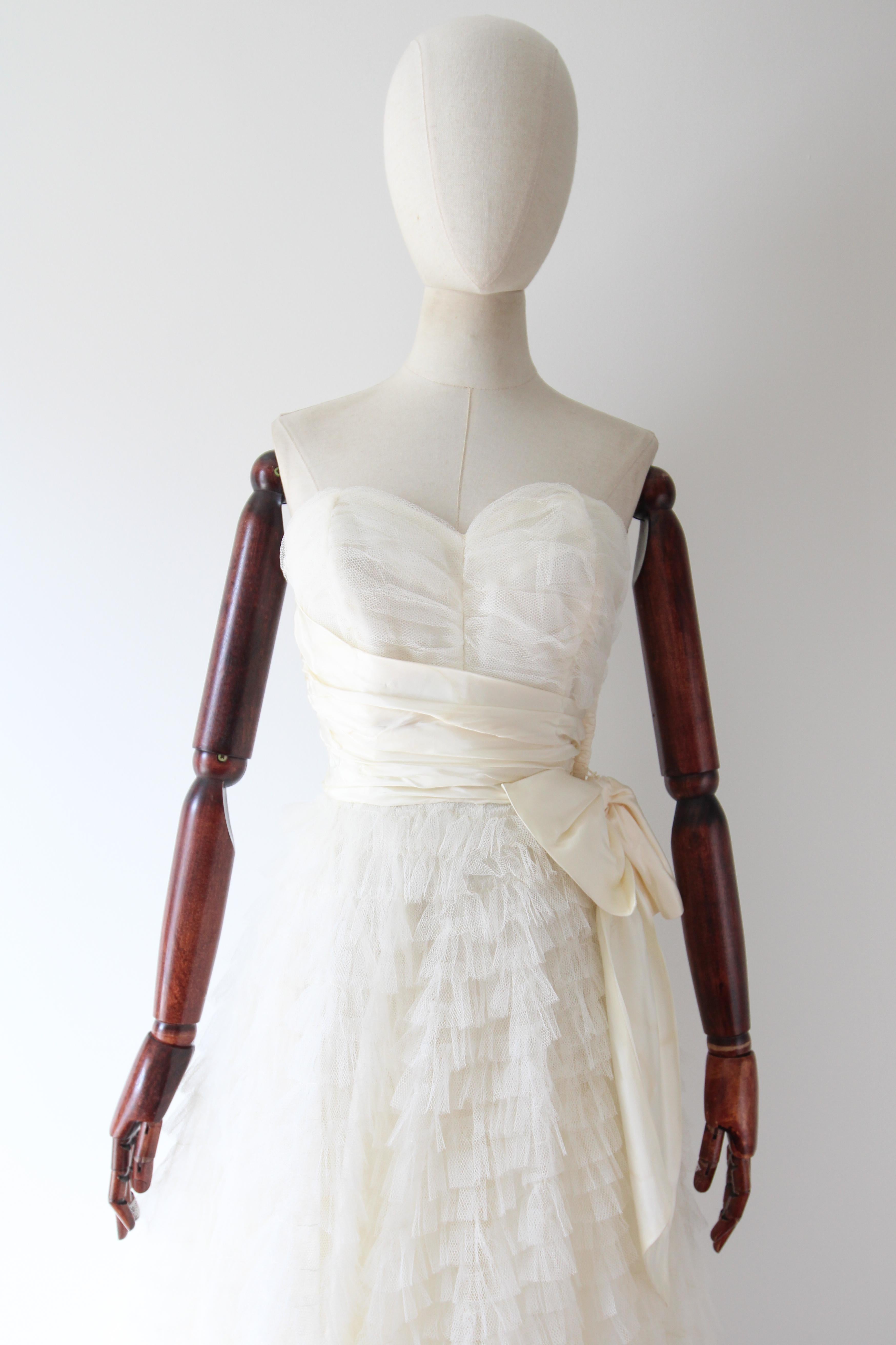 Dieses zeitlose und kultige Kleid aus den 1950er Jahren aus weißem, geschichtetem Tüll mit zarten, plissierten Viskose-Akzenten ist das perfekte Stück für einen besonderen Anlass.

Der trägerlose Ausschnitt des Kleides zeigt ein herzförmiges