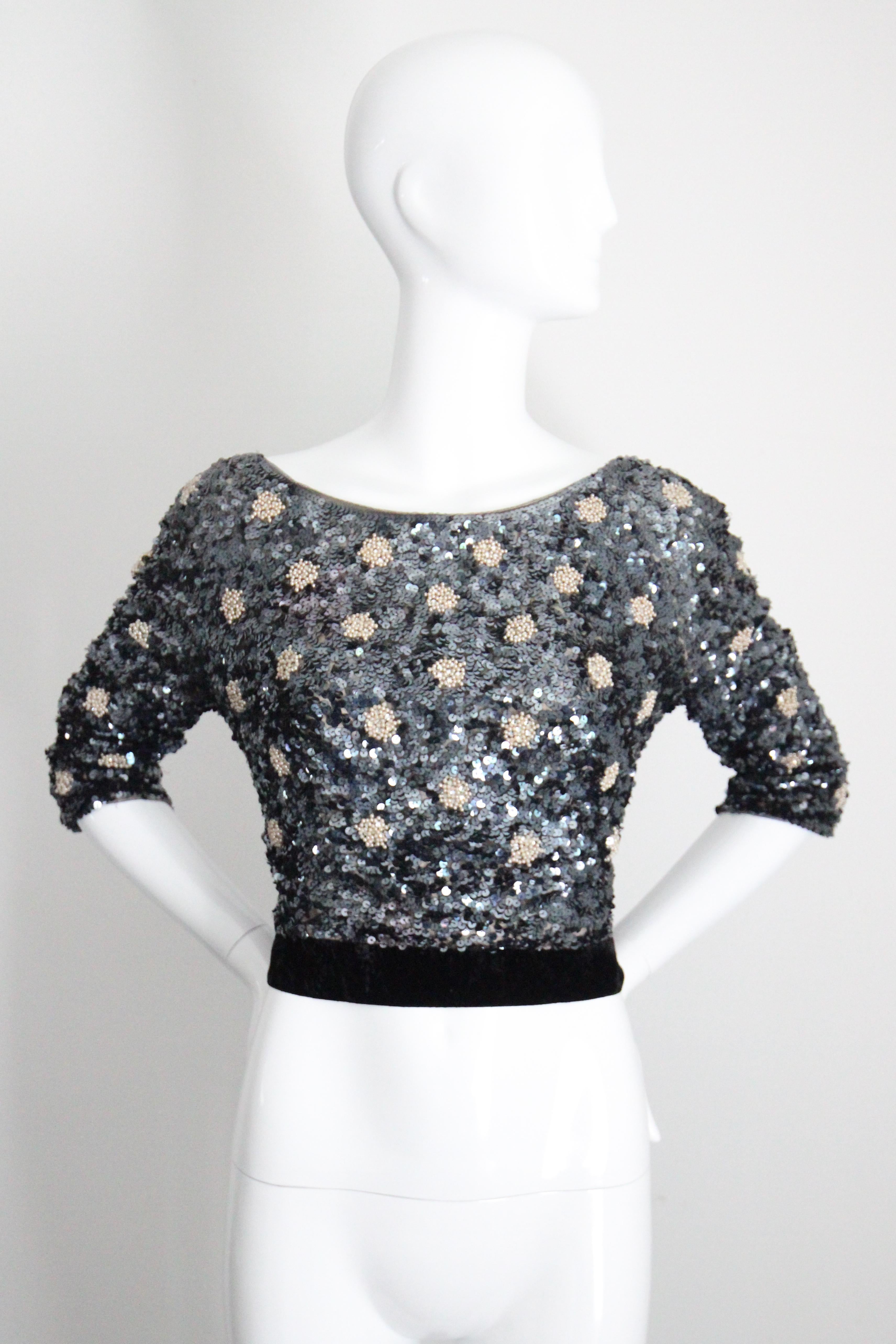 Rendered in a wonderful palette of steel blue sequins with scattered circular pearlescent beaded details surrounding silver claw set rhinestones, and set off by a black velvet detailed hemline, this original 1960's beaded blouse is the perfect touch
