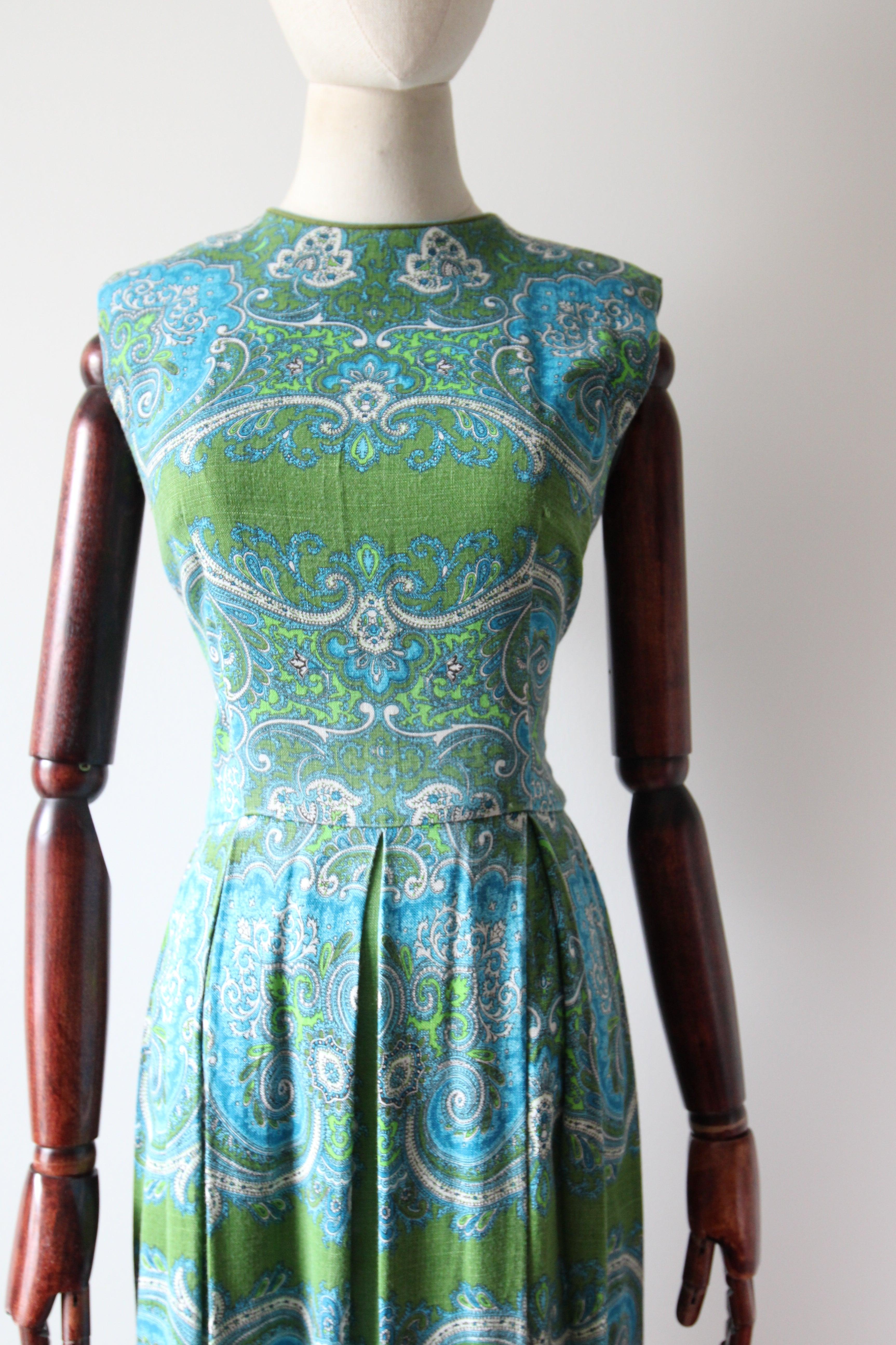This vibrant 1960's cotton dress, in a bright palette of greens and blues, with contrasting white accents in a paisley pattern, is just the piece to brighten up your seasonal wardrobe. 

The rounded neckline is framed by a border of green and