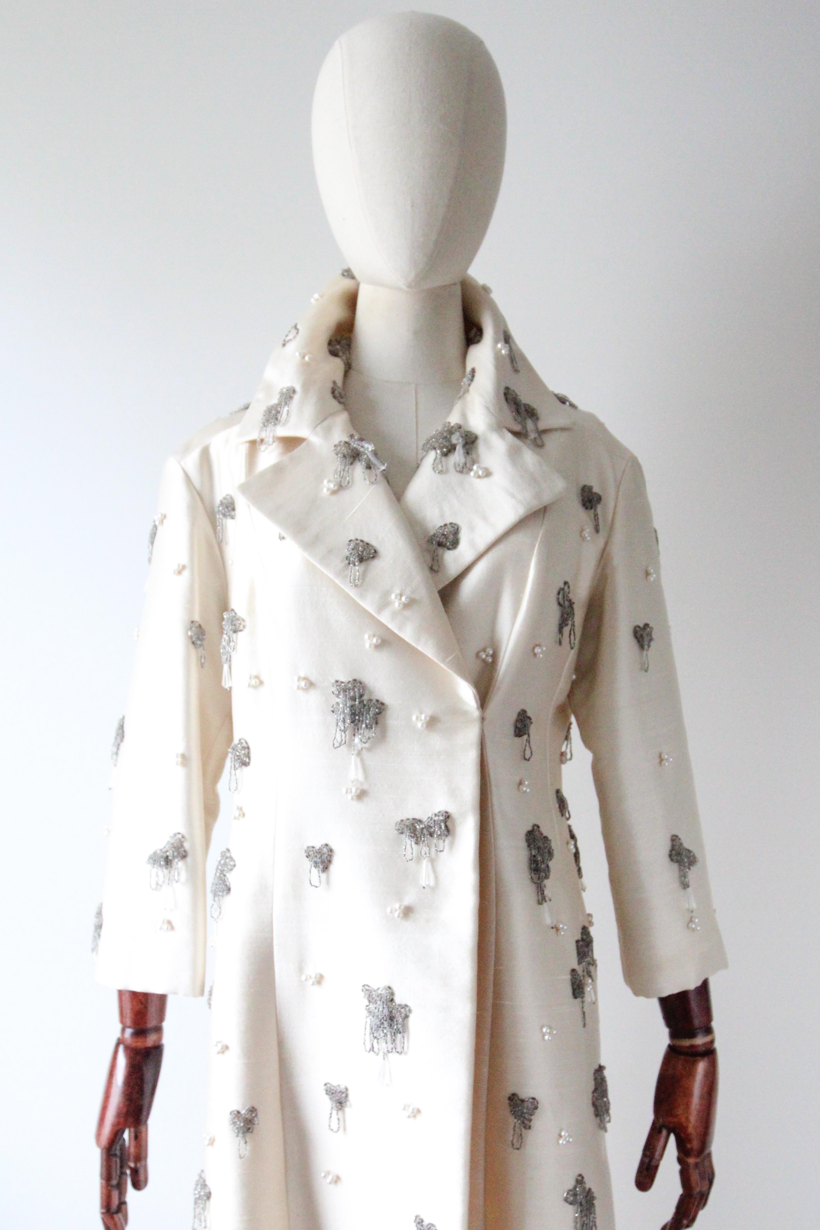 A beauty of her time, this original 1960's raw silk coat, rendered in cream coloured silk, which is in turn embellished with silver glass beads in draped and looped clusters, accented with faceted glass droplet beads, which are set off by clusters