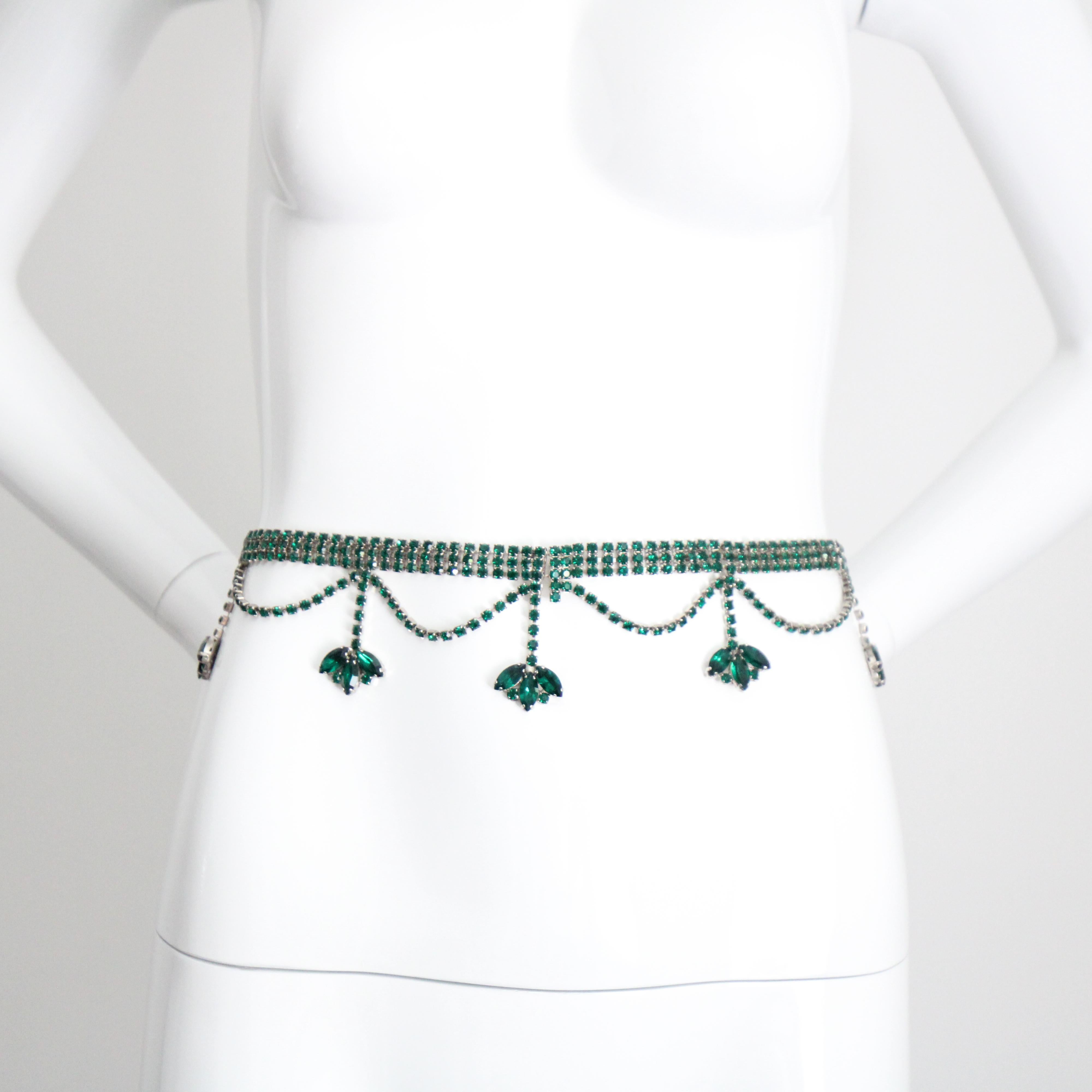 Questa spettacolare cintura anni '60, composta da strass di vetro color verde smeraldo con montatura in metallo argentato, è il pezzo forte perfetto per il tuo abbigliamento. La base a catena con triplo intarsio di strass si unisce centralmente