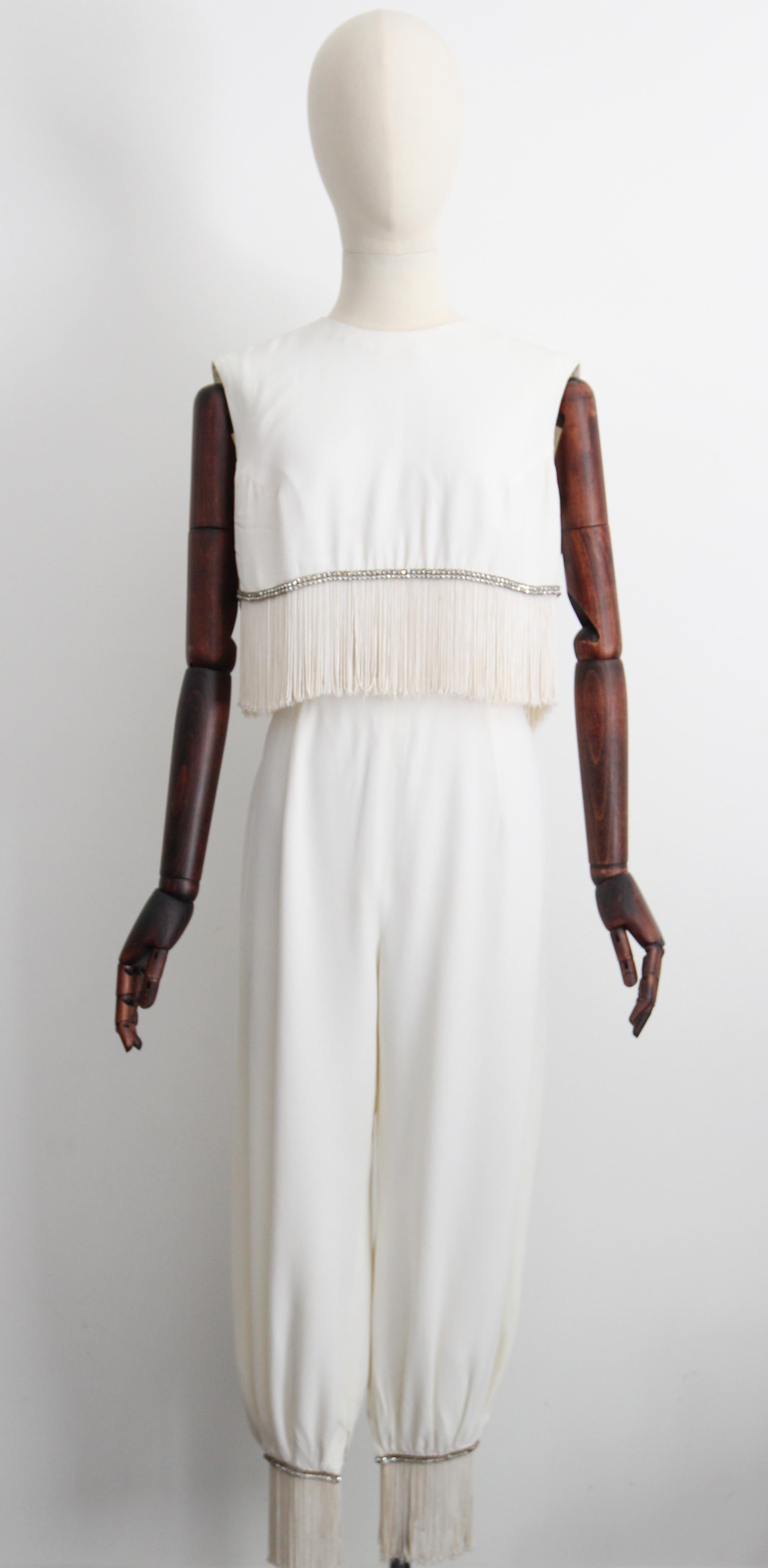 A piece to behold, this original 1960's ivory Lilli Diamond ensemble composed of a cropped blouse and matching high rise genie trousers, edged with strands of silver claw set rhinestones and cream fringing , is the perfect addition to your cocktail