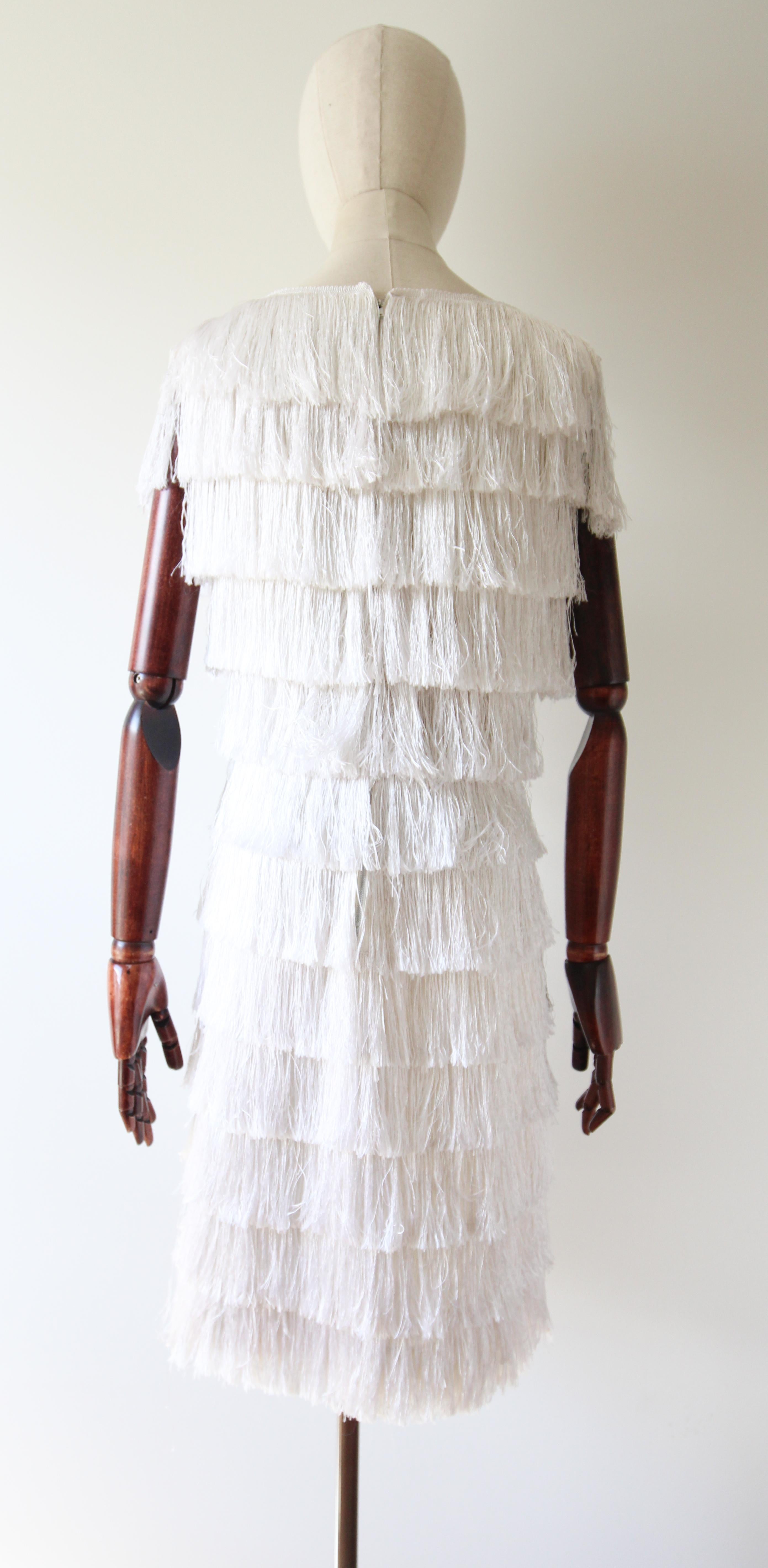 A vision in motion. This original 1960's ivory wiggle dress, accented by tiers of ivory fringing, upon a cream rayon base, is just the piece to add to your vintage collection. 

The bateau neckline of the dress is framed by scalloped ivory braiding,