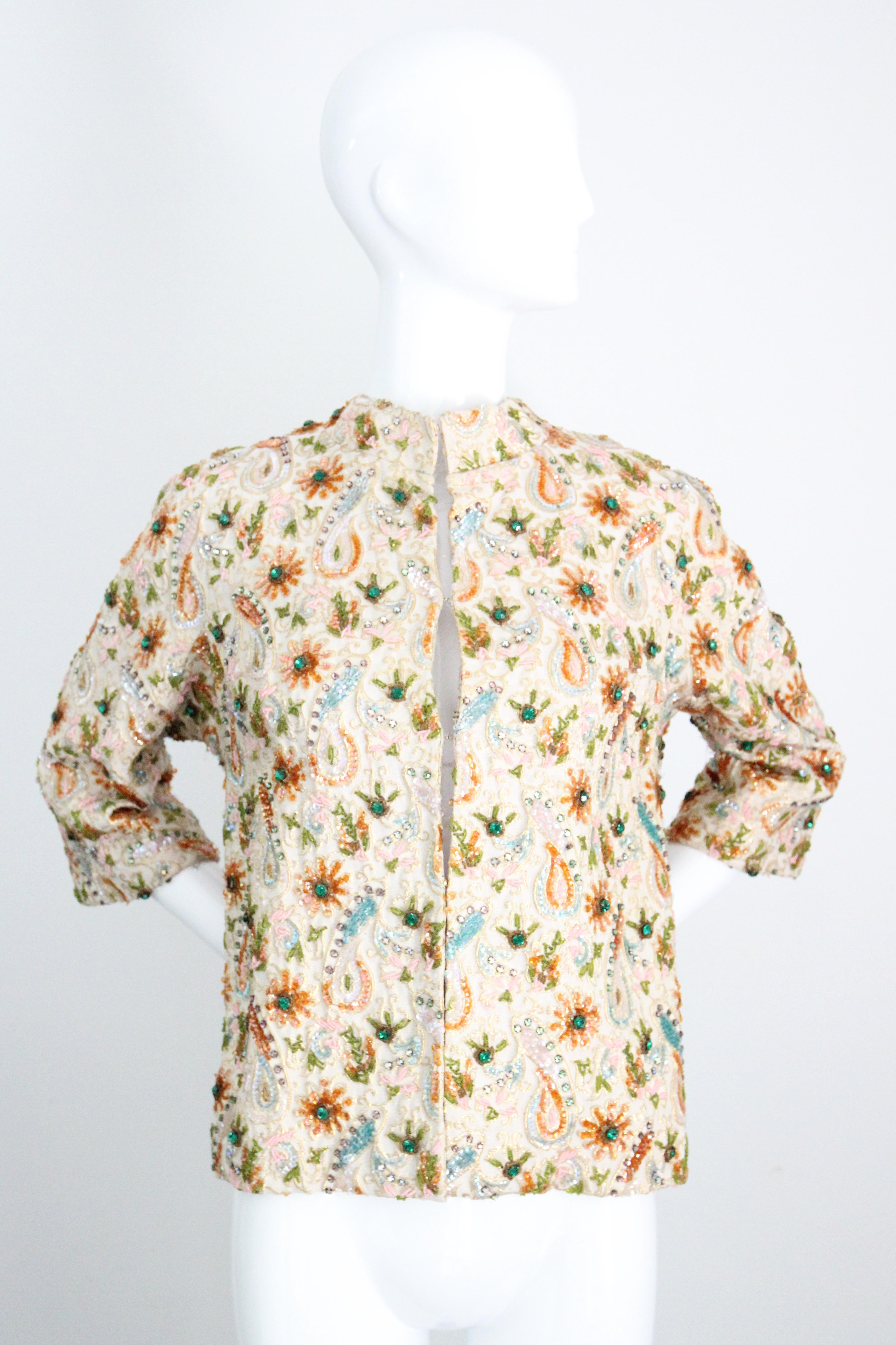 This truly mesmerising 1960's jacket, rendered in a cream and gold lurex brocade, which is embellished with sequins in shades of iridescent amber, pale blue, turquoise blue and clear sequins, accented by glass beaded accents in shades of pink, green