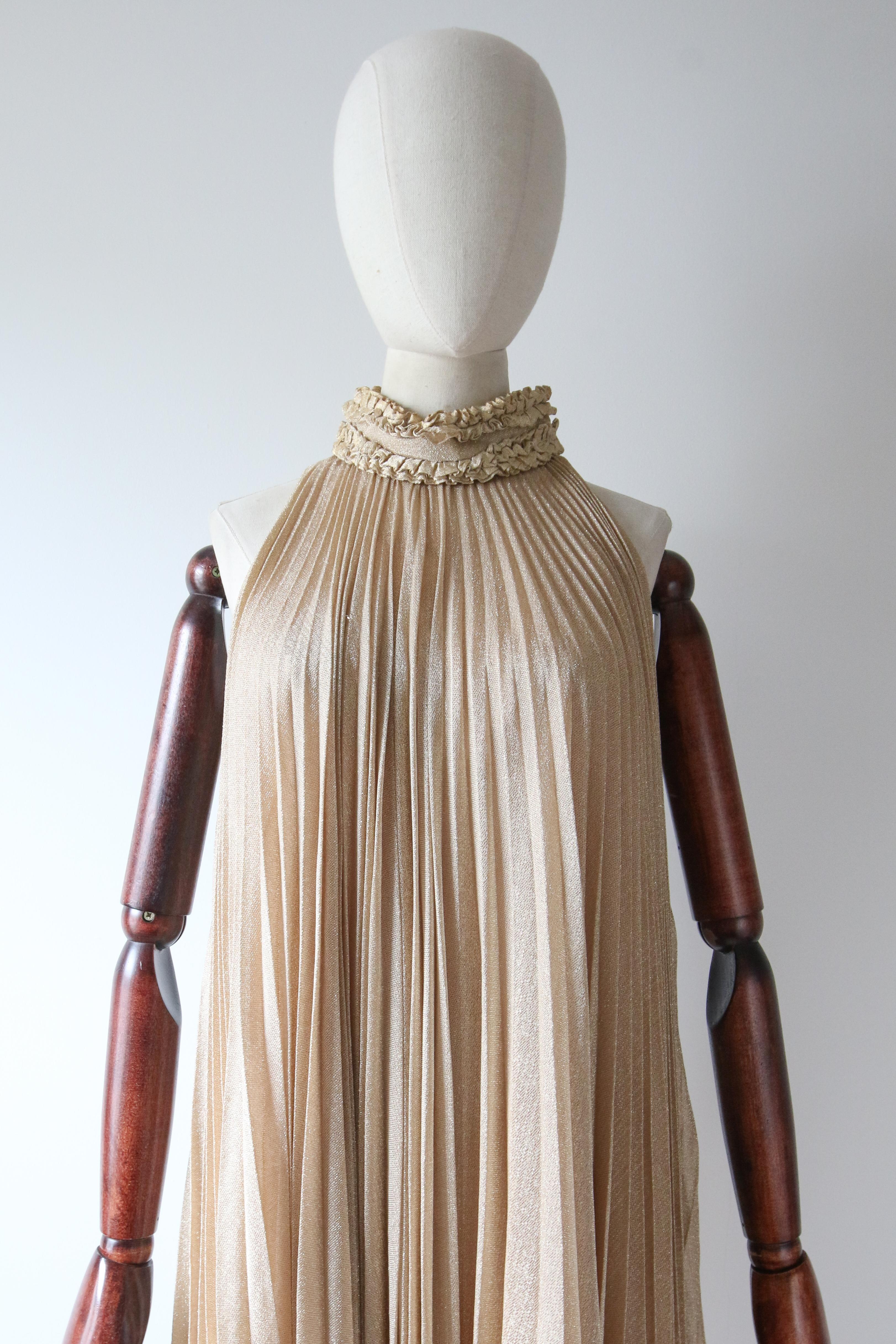 Este vestido original de lúrex dorado de los años 60, con pliegues en acordeón y trenzado dorado a lo largo del escote, es una auténtica pieza de la historia de la moda. 

El escote de cuello alto se abrocha en el lado izquierdo con botones a