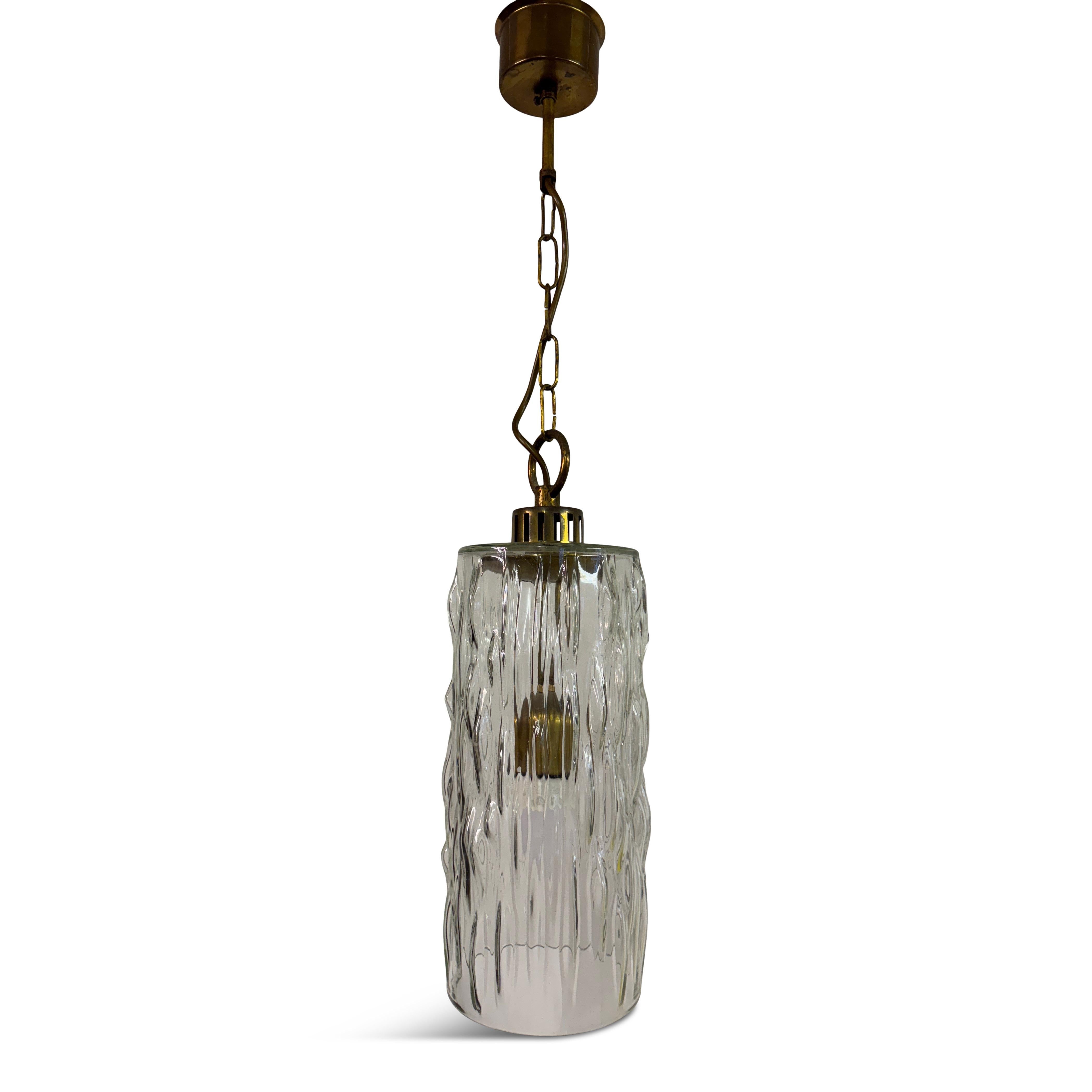 Una lampada a sospensione belga degli anni '60 con un paralume in vetro profondamente strutturato sospeso da una catena e un rosone in ottone patinato. 

La forma cilindrica è semplice, ma il vetro spesso e modellato aggiunge un effetto scultoreo,