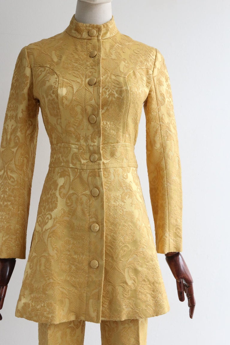 This truly iconic 1960's trouser suit by Janice Wainwright for Simon Massey, in a vibrant gold and yellow brocade, is the perfect ensemble to add to your iconic wardrobe.

The mandarin collar of the jacket is framed by a simple shoulder line. The