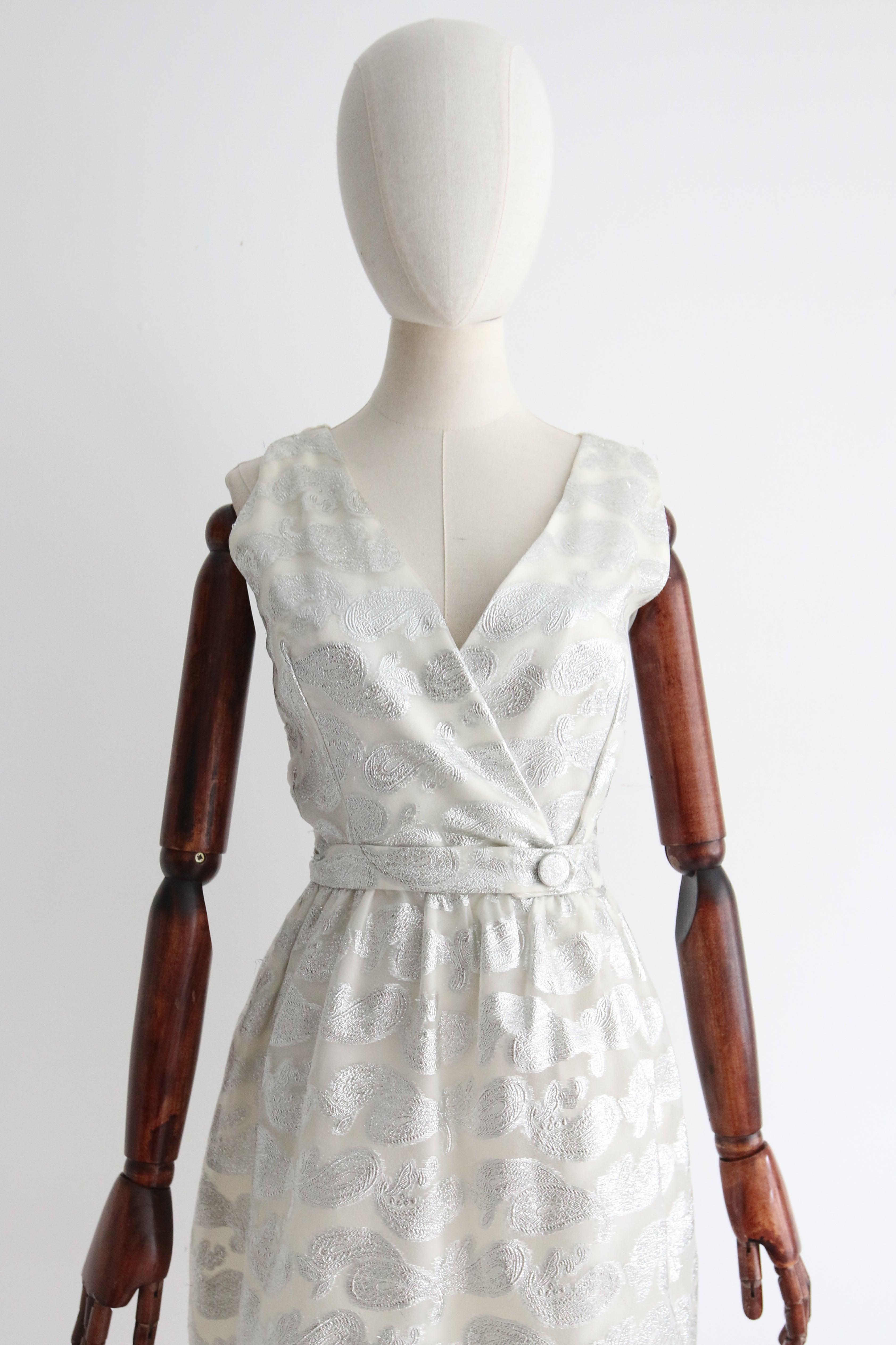 Este brillante vestido de los años 60 de lúrex plateado y brocado de organza blanca con estampado paisley,  es el pequeño número perfecto para añadir un toque de brillo a tu vestuario. 

El profundo escote en V se acentúa con el diseño cruzado del