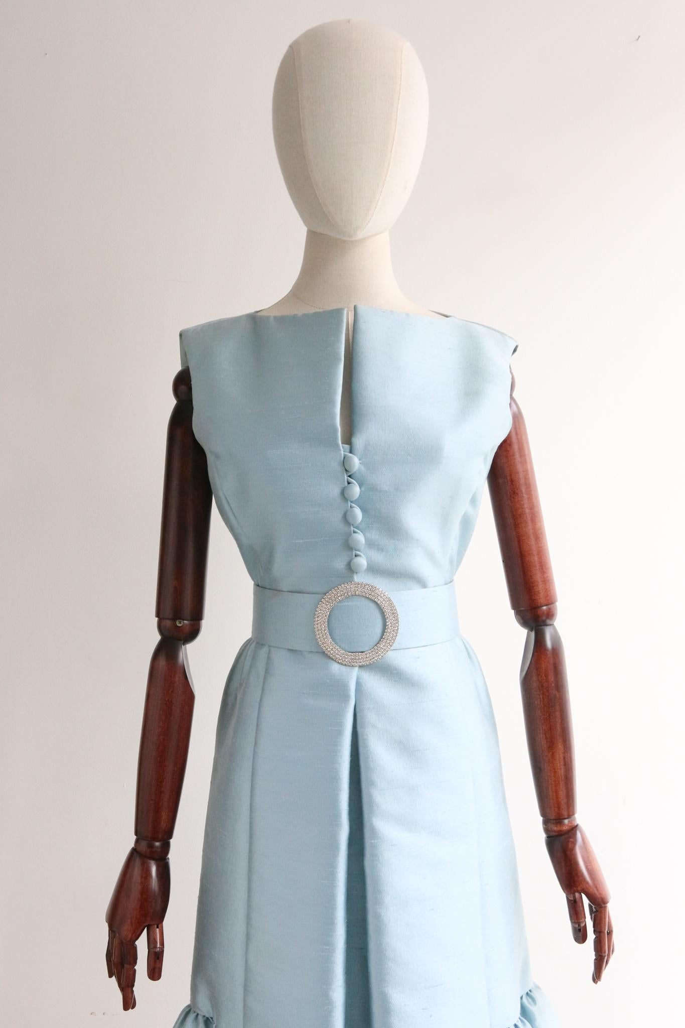 Made in the most iconic shade of ice blue shantung silk, this mesmerising 1960's Teal Traina dress is a rare piece to behold. 
The straight cut bateau neckline boasts a central split along the décolletage and is framed by a sleeveless cut. The